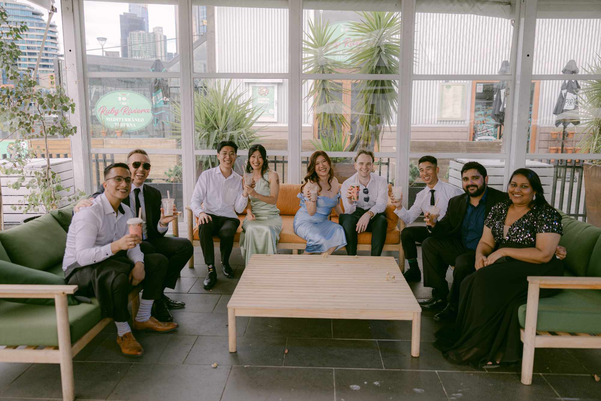 Wedding guests sit together on lounge sofas, smiling and holding drinks in a bright indoor venue.