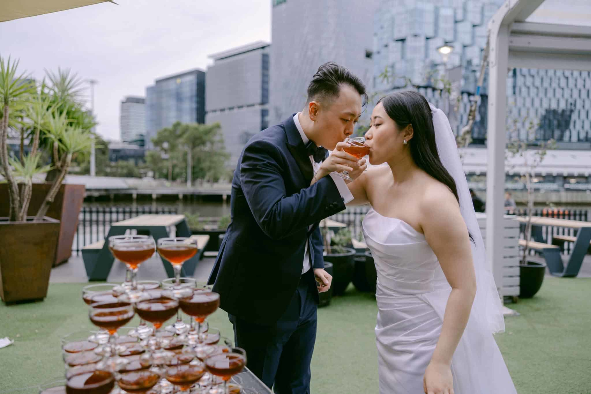Bride and groom share a toast beside a champagne tower at an outdoor city wedding venue.