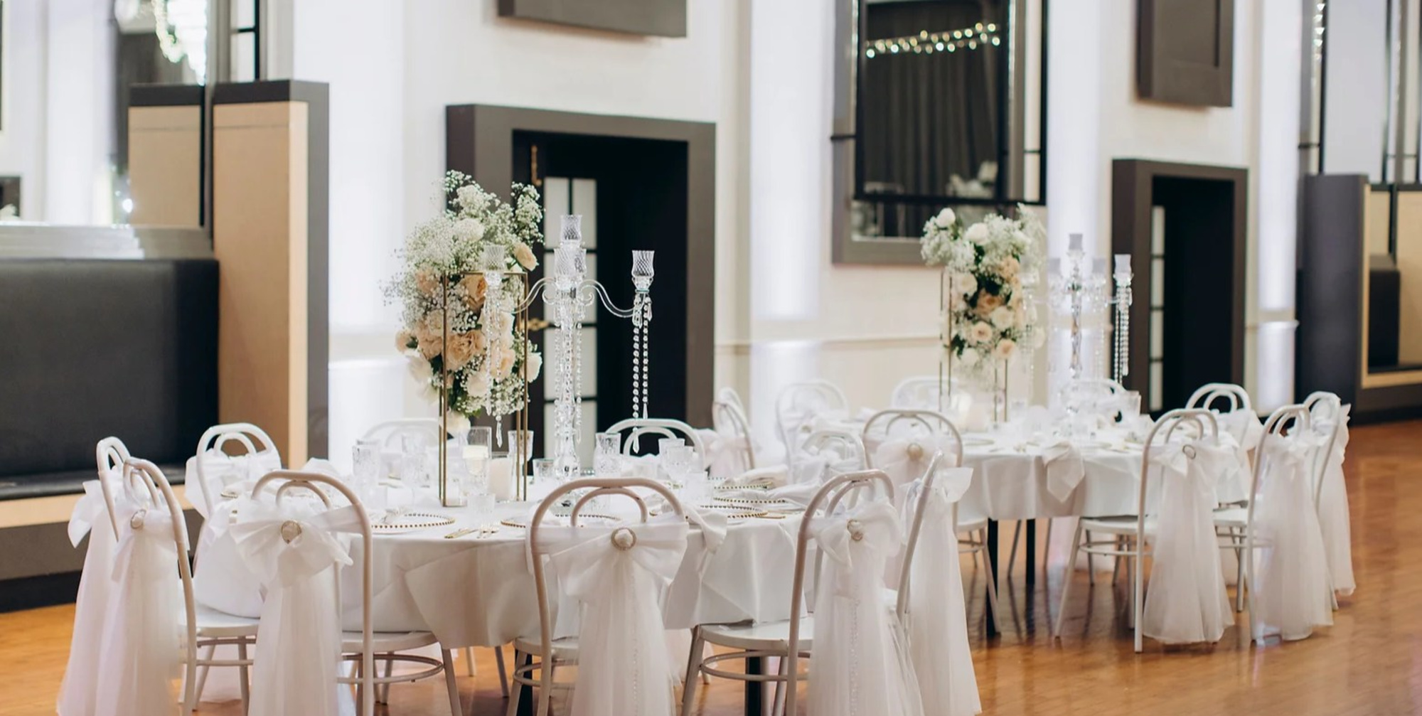 Elegant indoor wedding reception with round tables, white chairs, and tall floral and crystal centerpieces.
