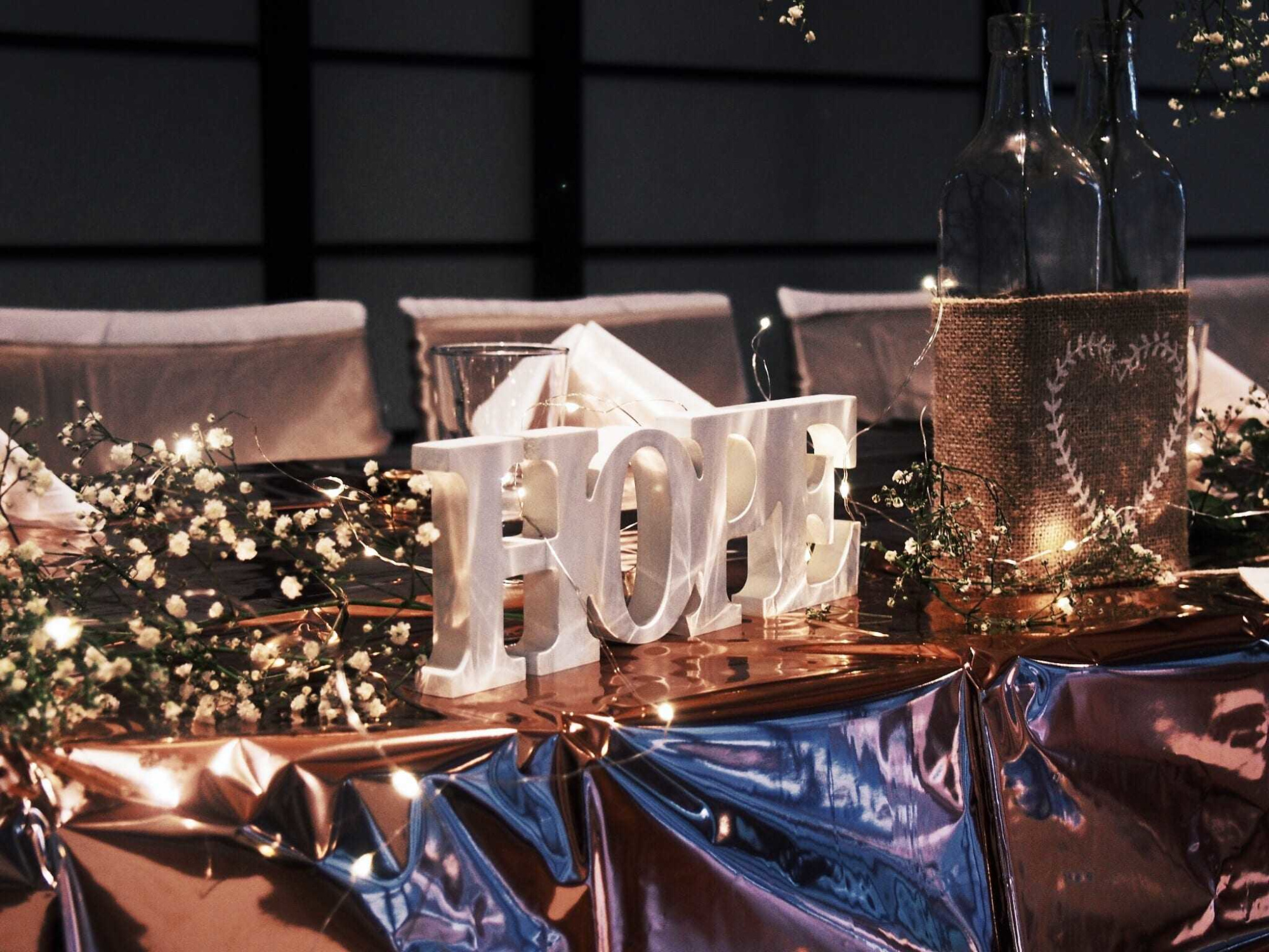 Rustic wedding table centerpiece with HOPE letters, glass bottles, burlap, fairy lights, and baby's breath on a metallic cloth.