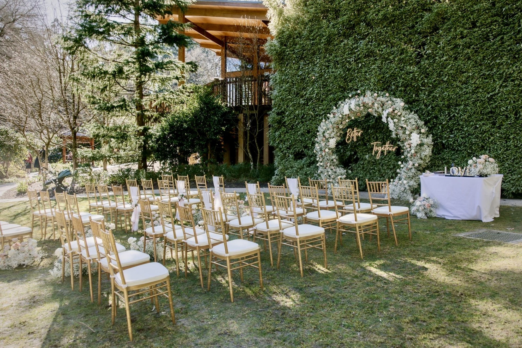 Outdoor garden wedding ceremony setup with gold chairs facing a large circular floral arch and greenery backdrop.