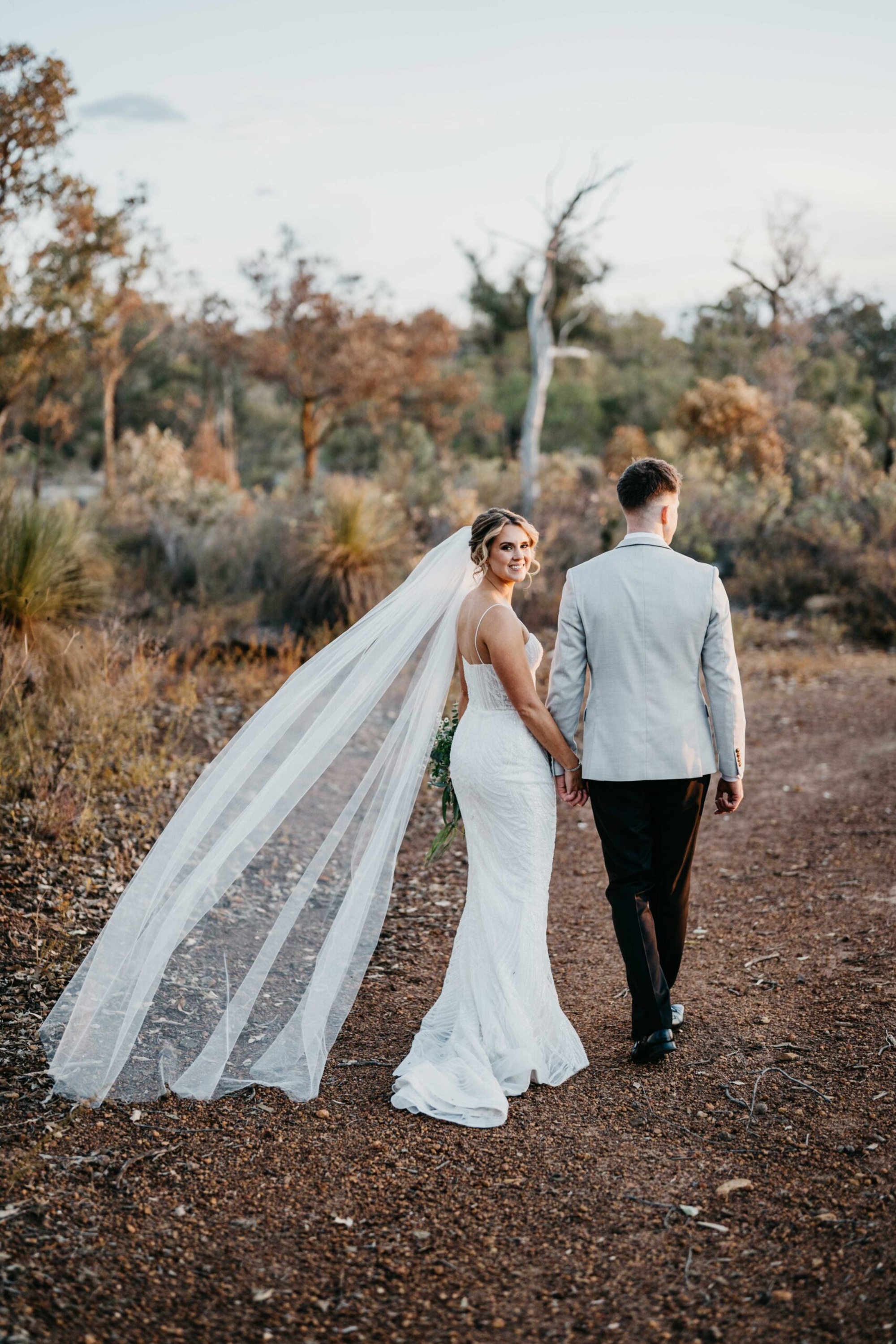 Bride and groom walk hand in hand along a rustic outdoor path, her long veil flowing behind them.