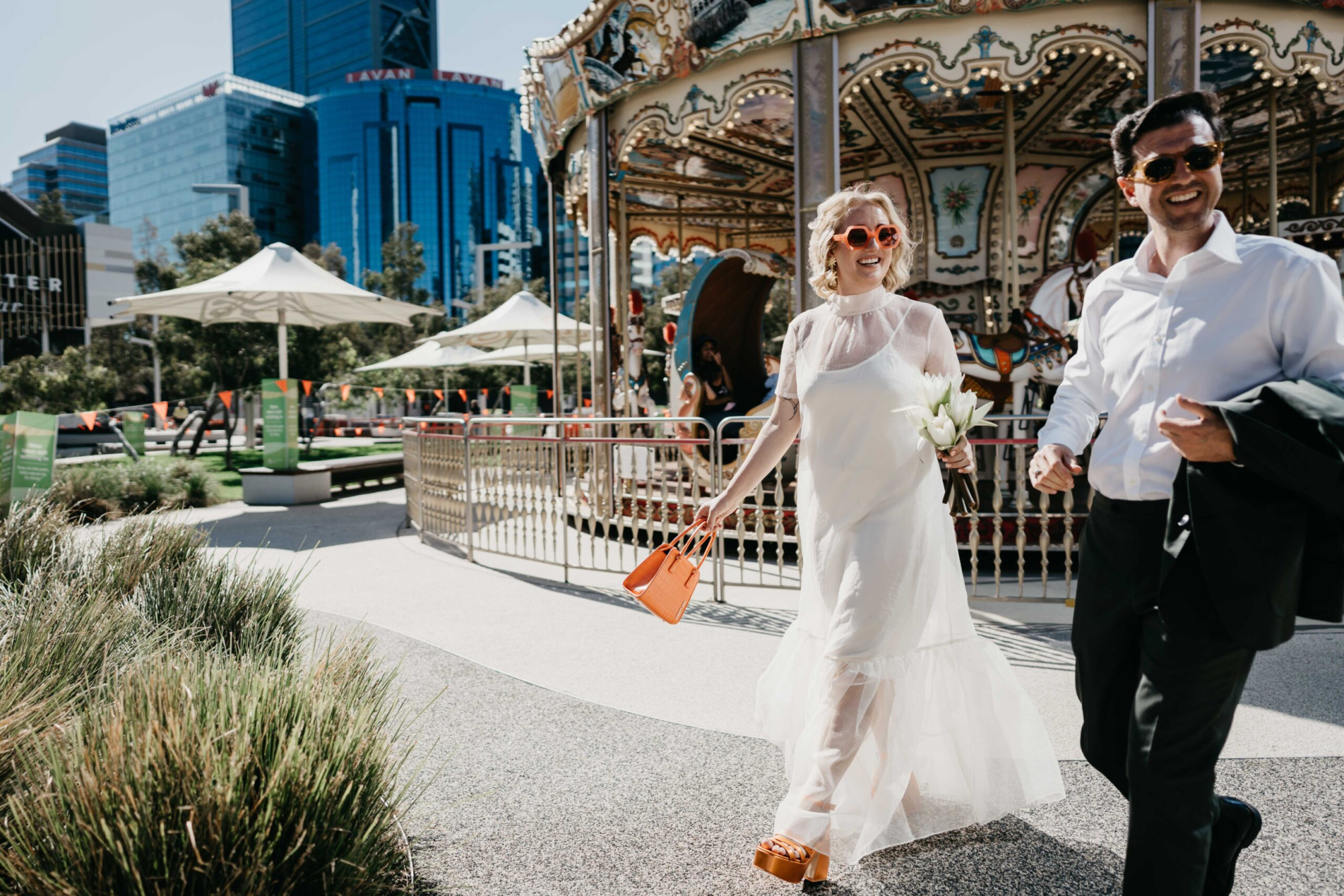Bride and groom walk joyfully past a carousel in a sunny urban park holding flowers and a bright orange bag.