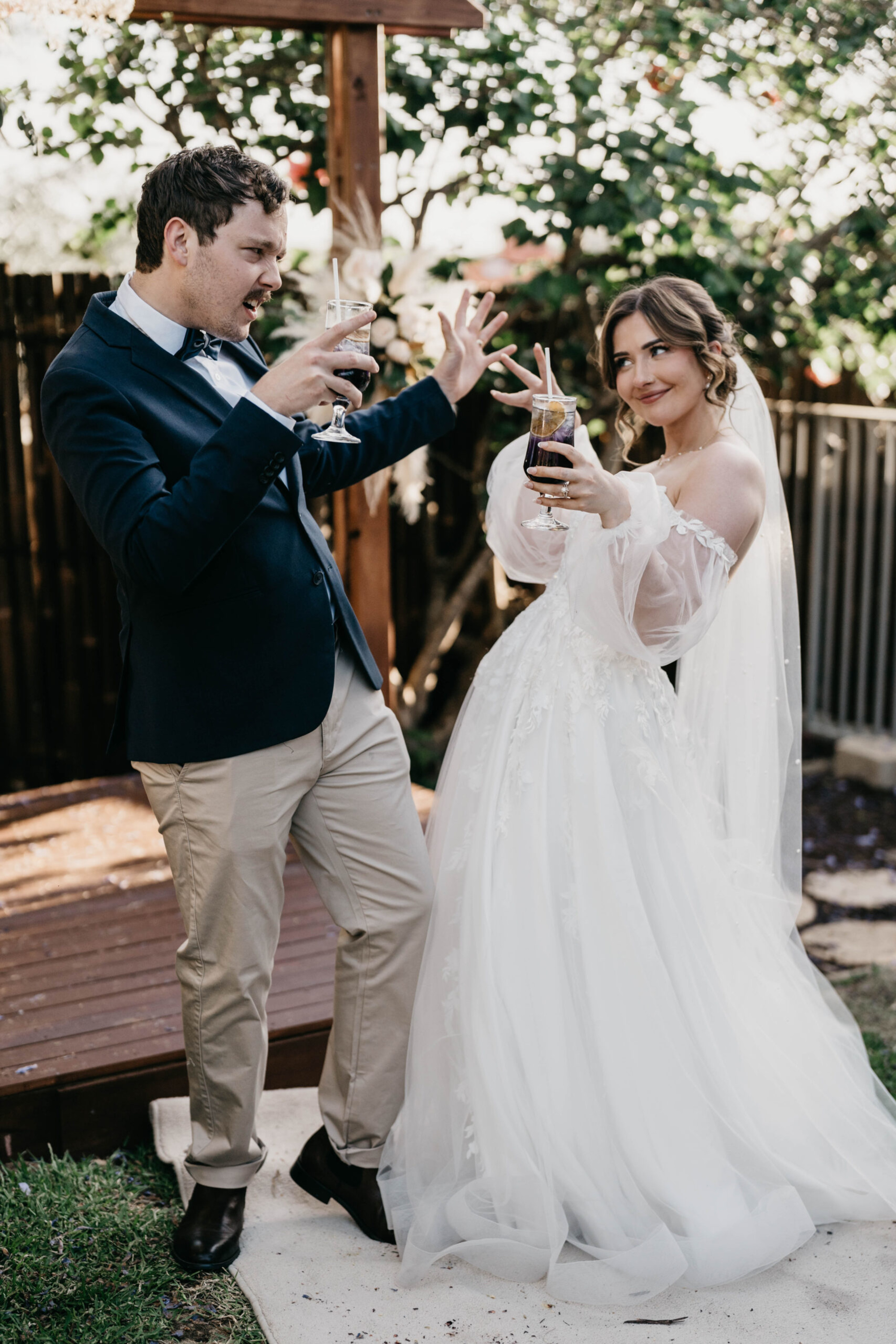 Candid outdoor shot of a bride and groom laughing and posing with drinks at their wedding.