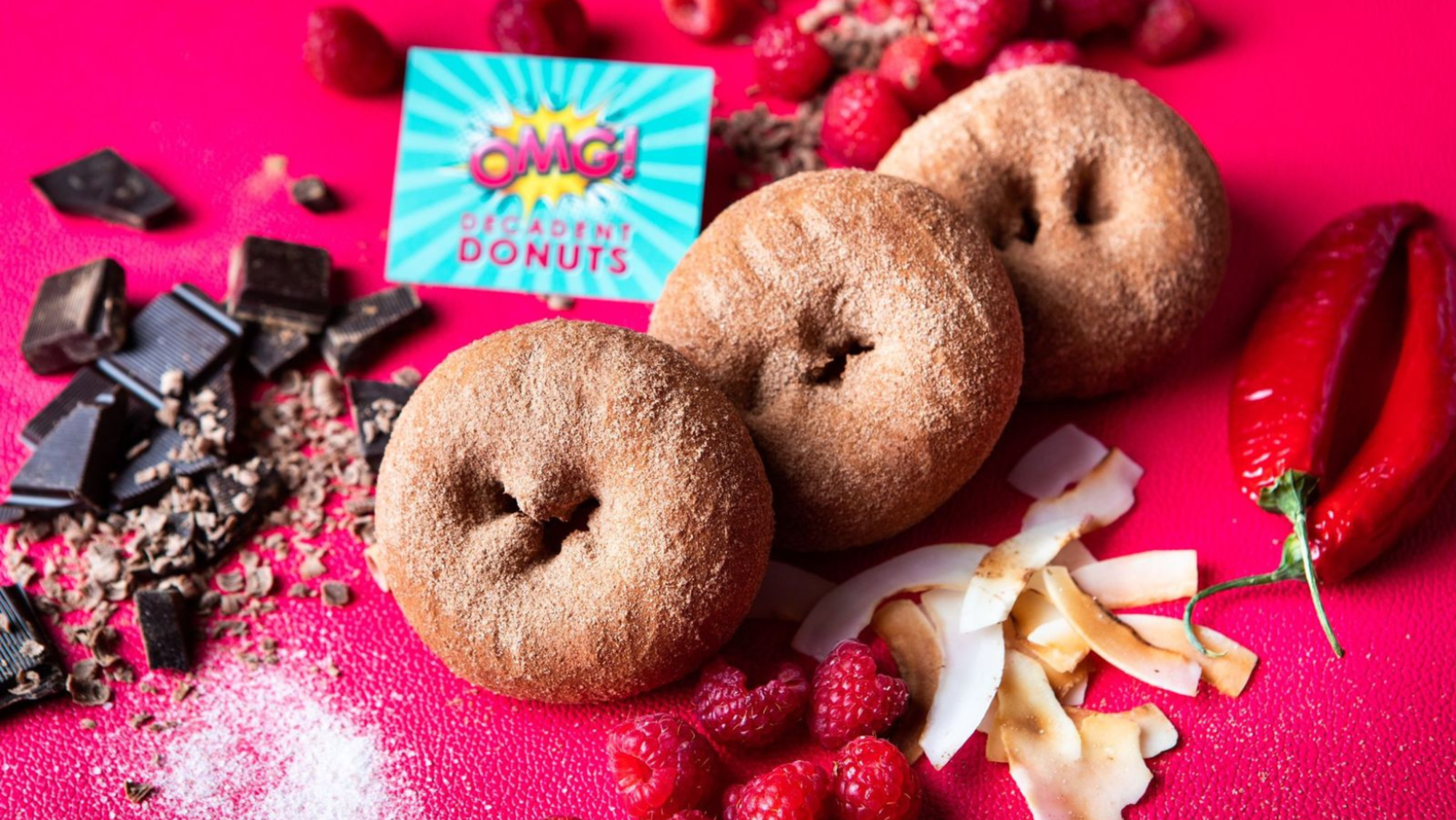 Cinnamon sugar donuts styled with chocolate, raspberries, coconut and chili on a bright pink background.