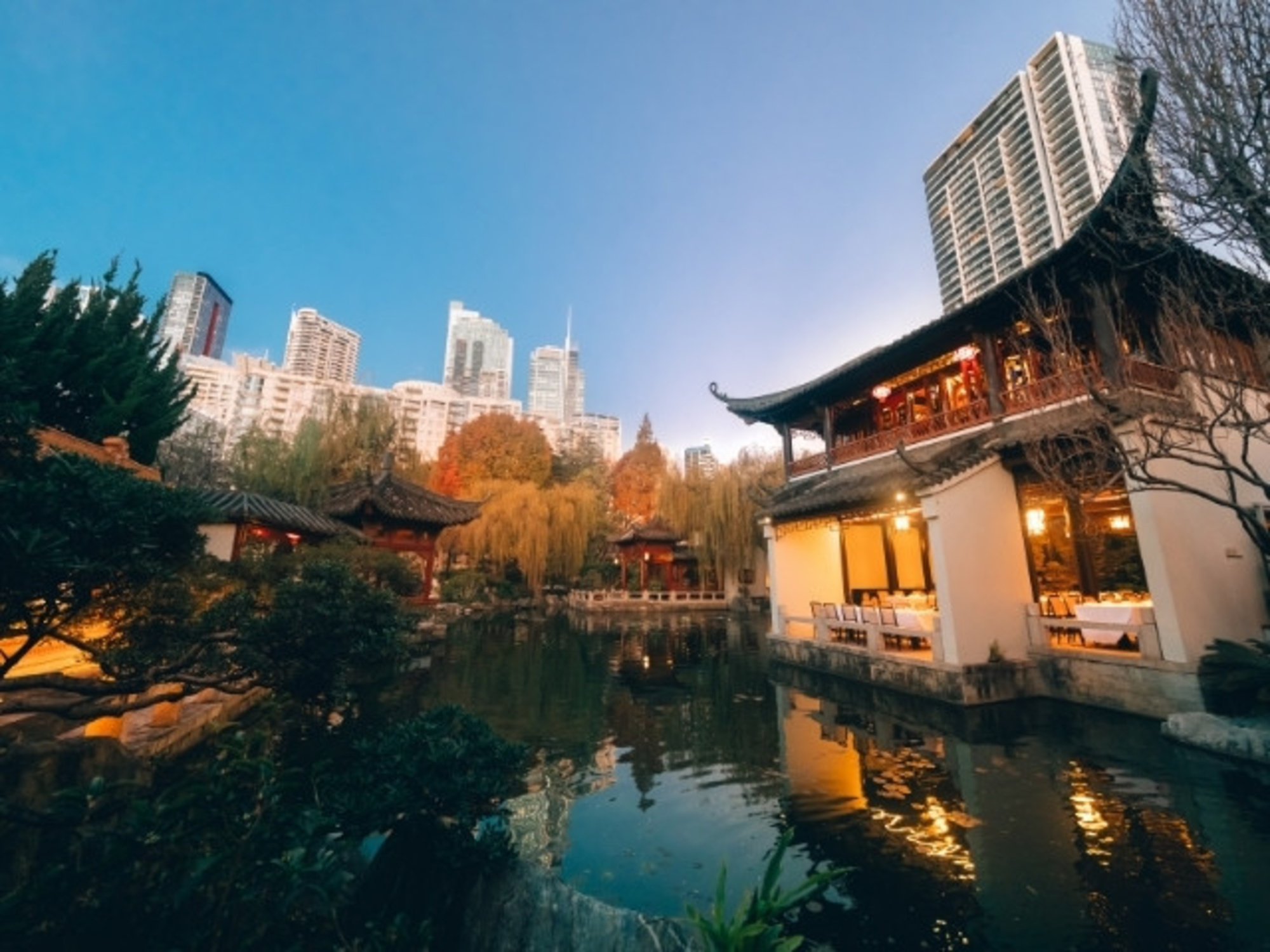 Tranquil Asian-inspired garden venue with ponds and traditional pavilions set against a modern city skyline at dusk.