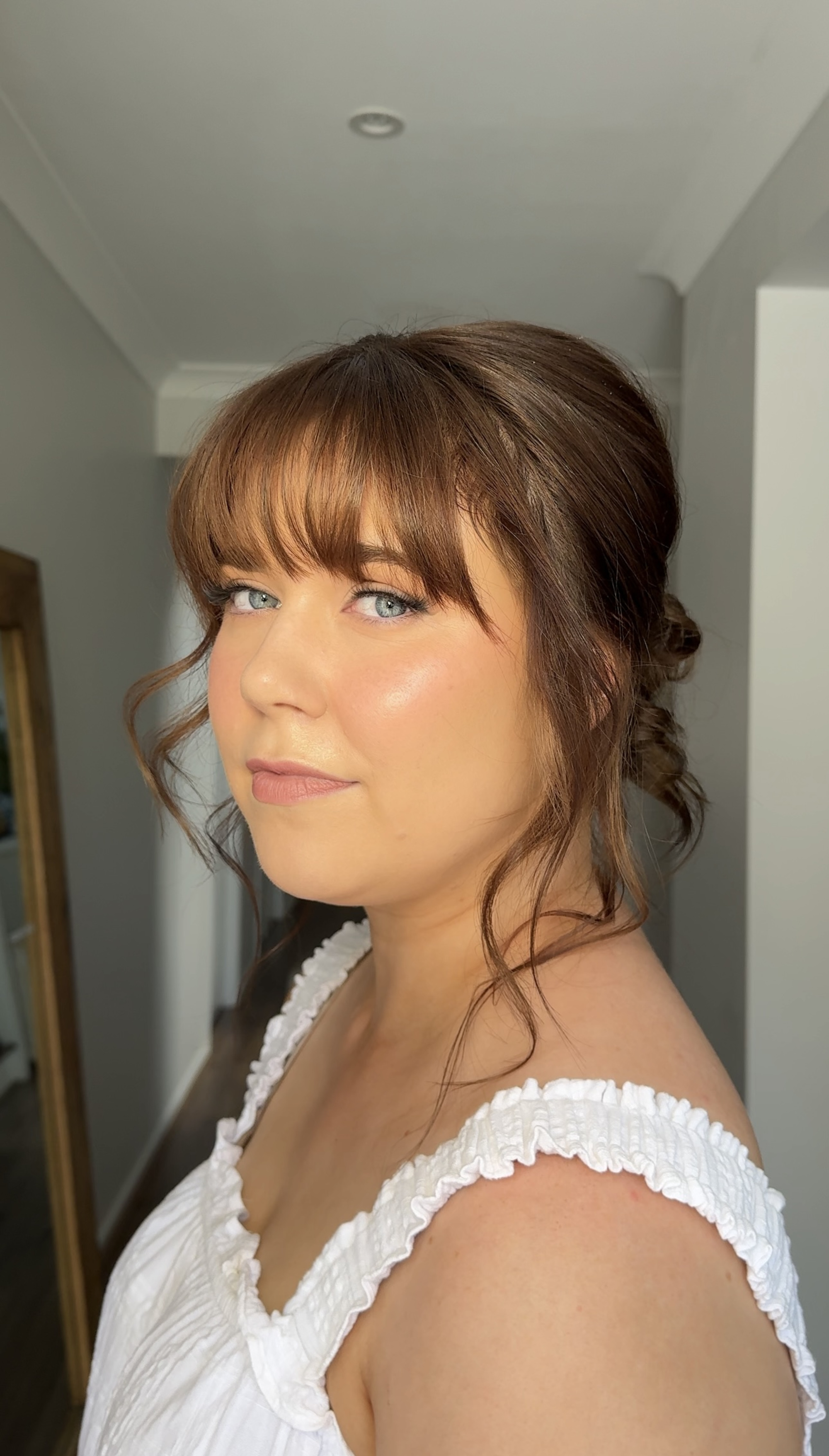 Woman with soft glam makeup and loose updo hairstyle posing in natural indoor light.