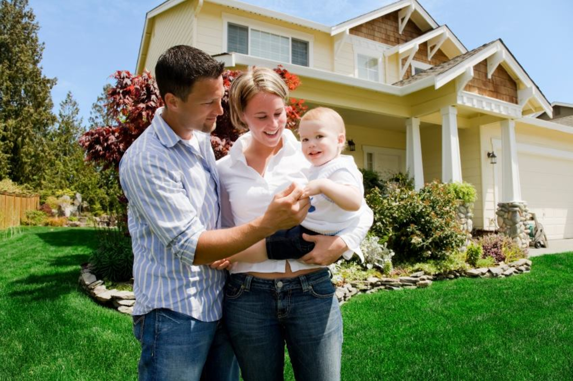 Smiling couple holds their baby while standing together in the front yard of a suburban home.