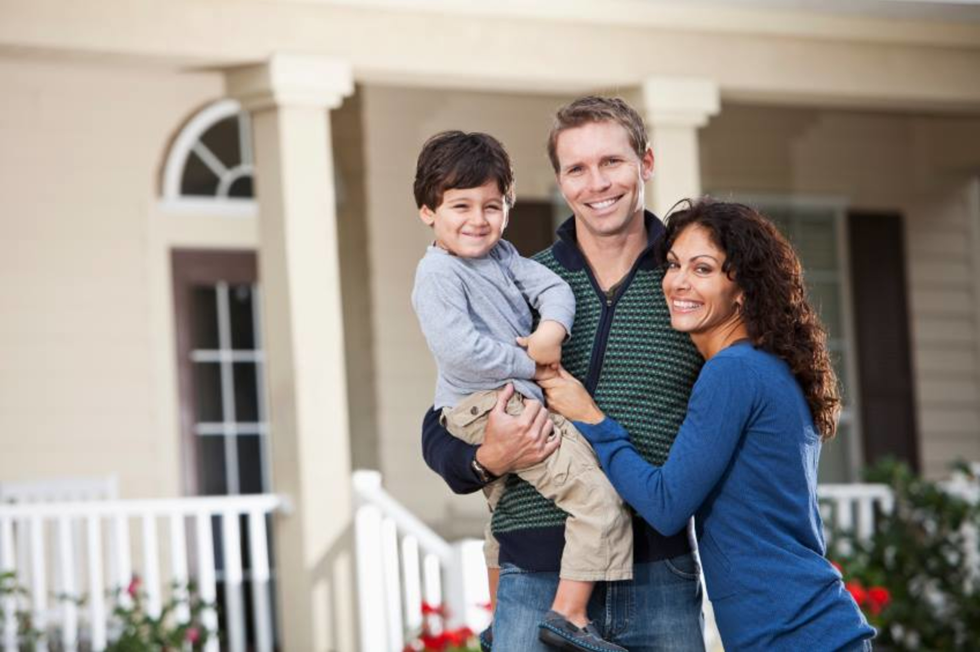Smiling family with parents holding their young child while posing together on a front porch.