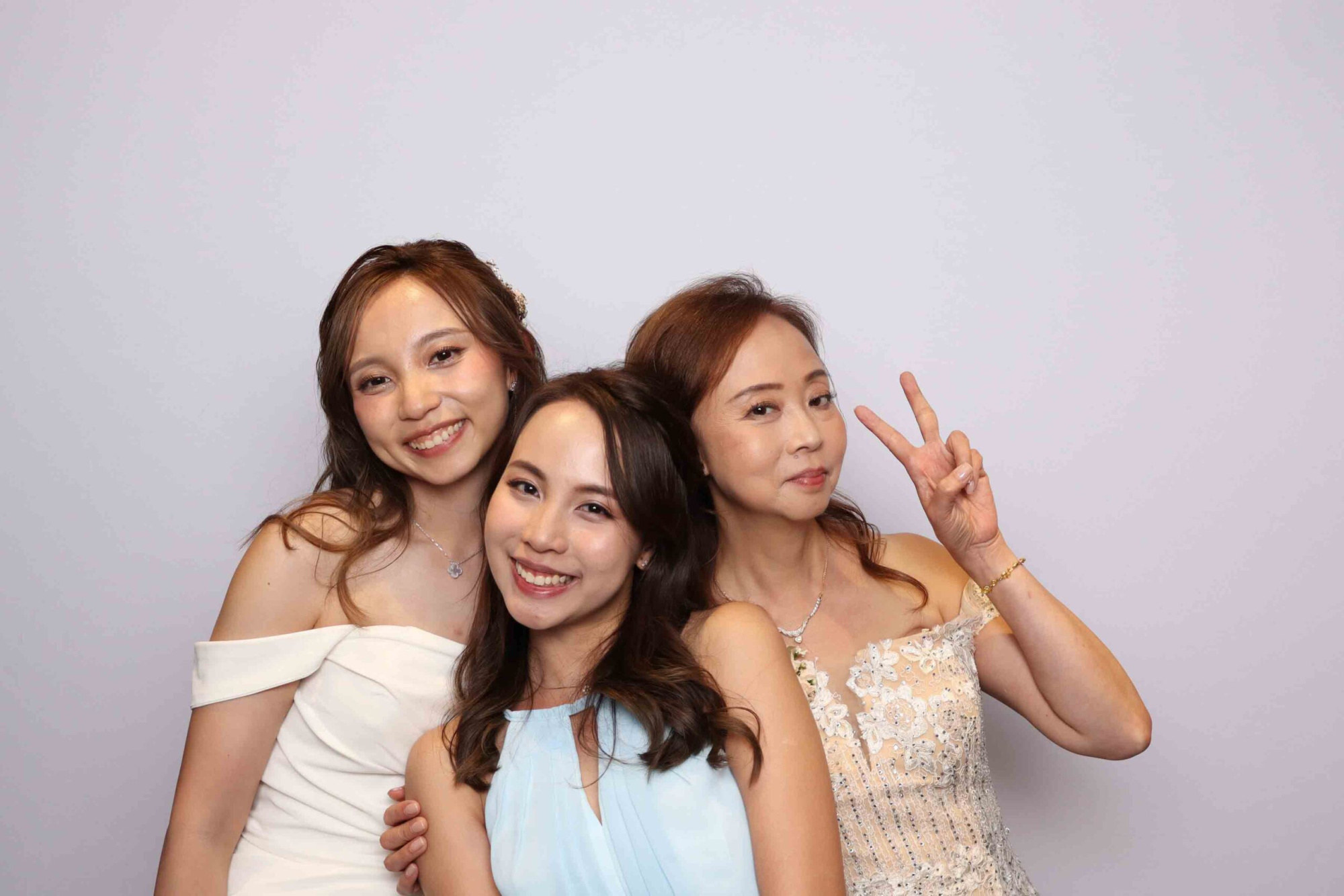 Three women in formal dresses smile and pose together against a simple light backdrop at a wedding.