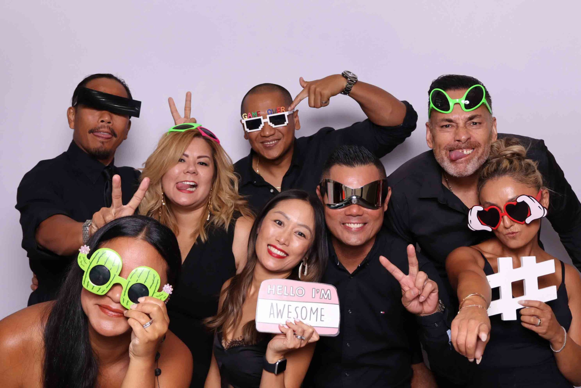 Group of wedding guests posing in a photo booth with playful glasses and props against a light backdrop.