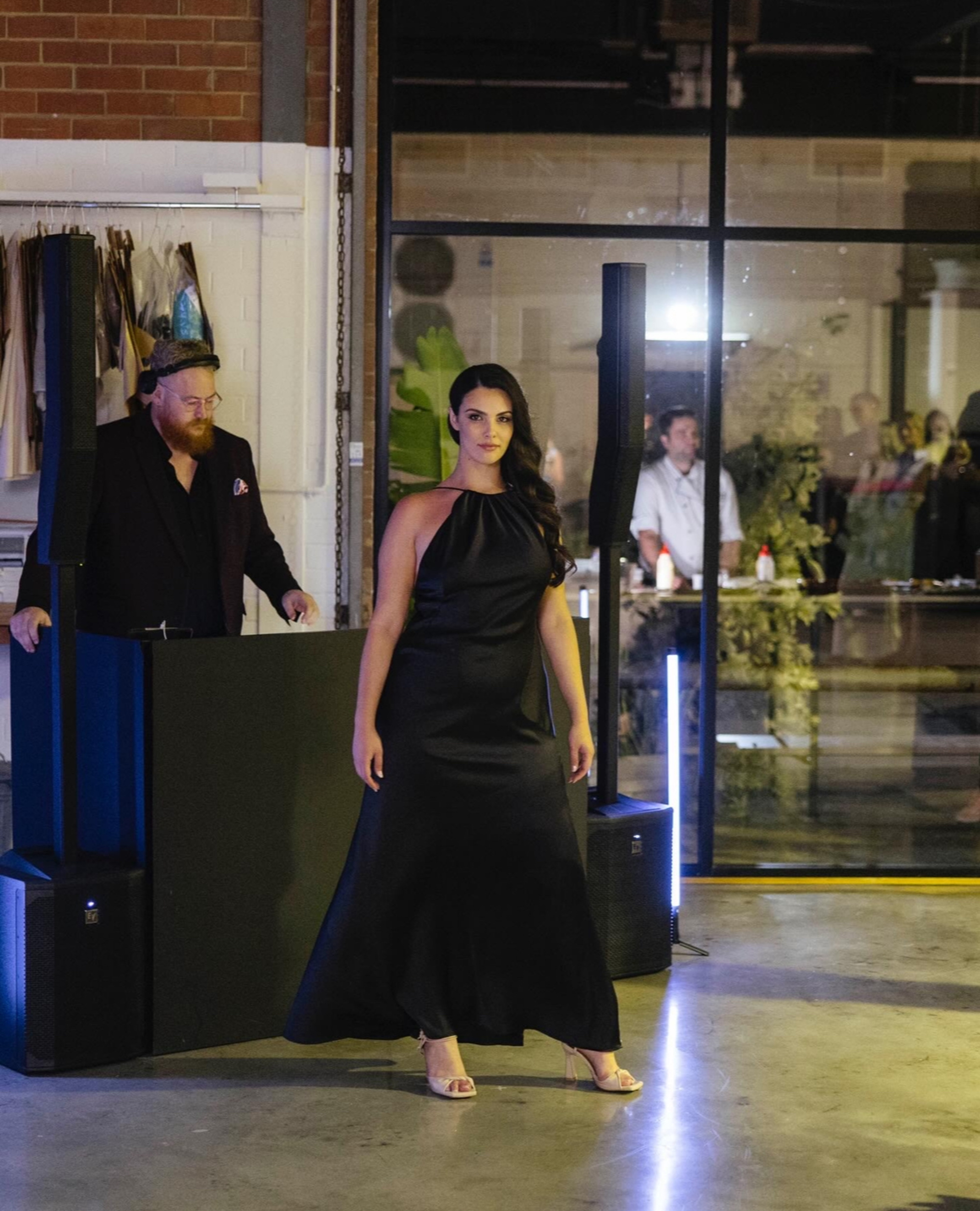 Woman in a sleek black gown poses on the dance floor in front of a DJ at an indoor wedding reception.