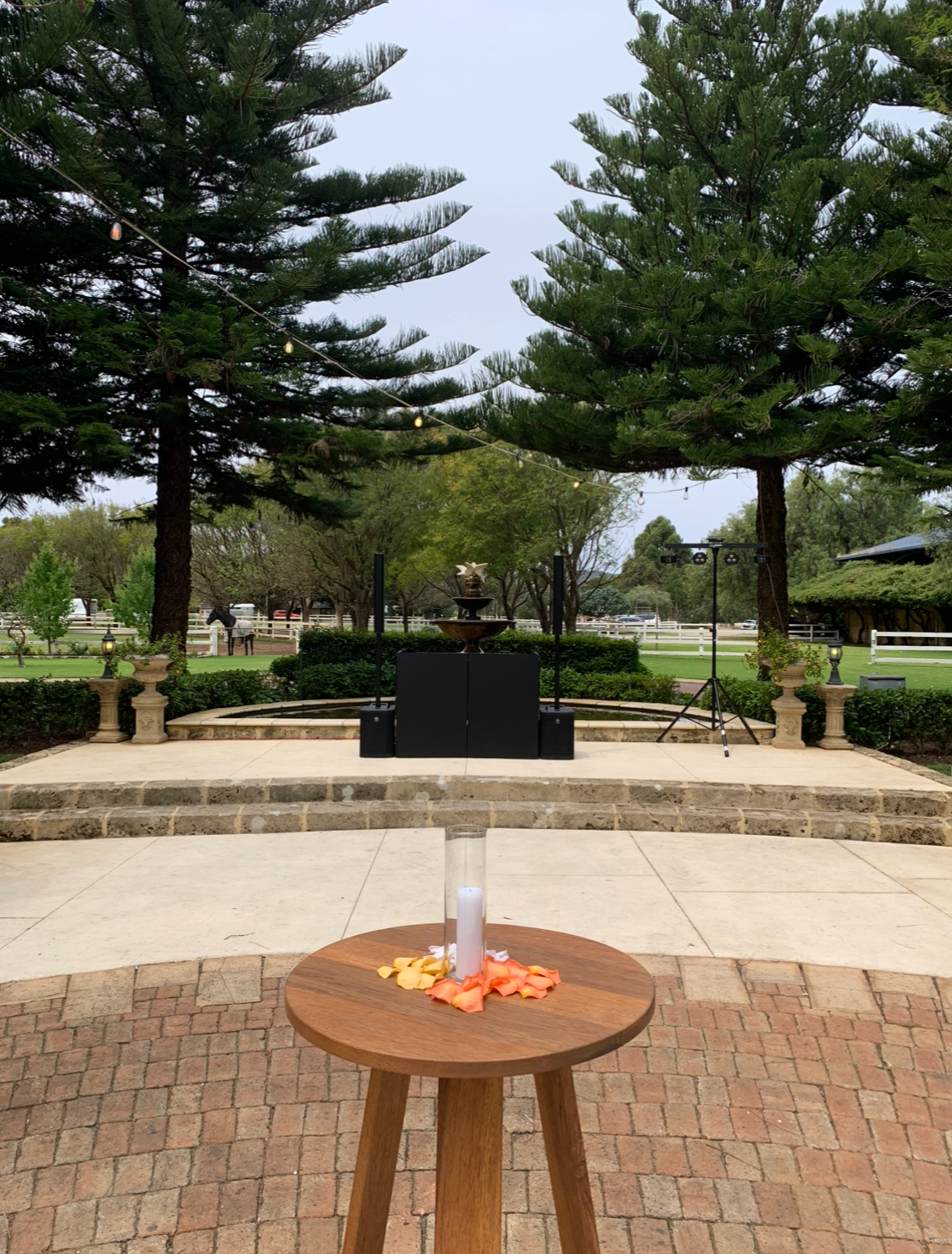 Outdoor wedding ceremony setup with a unity candle table in front of a garden stage and tall trees.