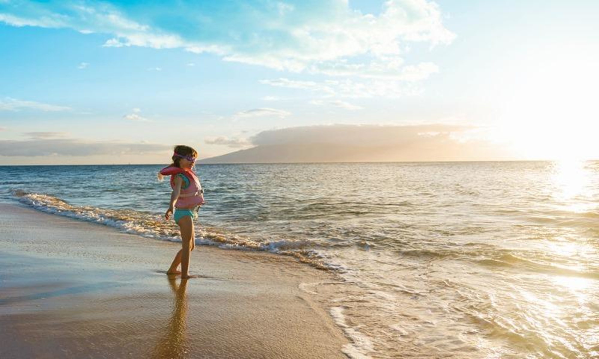 Child standing at the shoreline of a sunny beach with gentle waves and a distant island view.