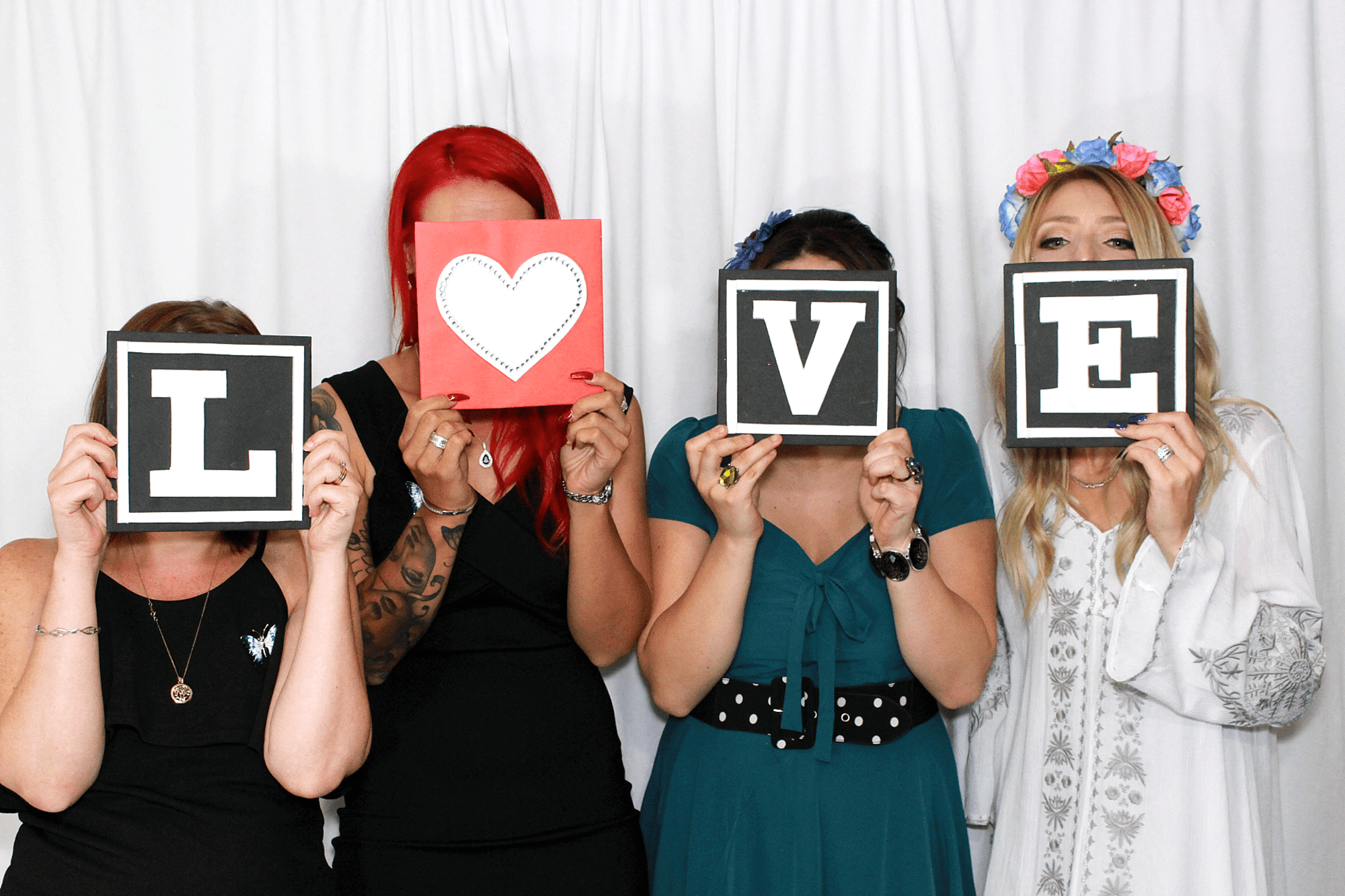 Four women in a photo booth hold letter cards spelling LOVE in front of their faces.
