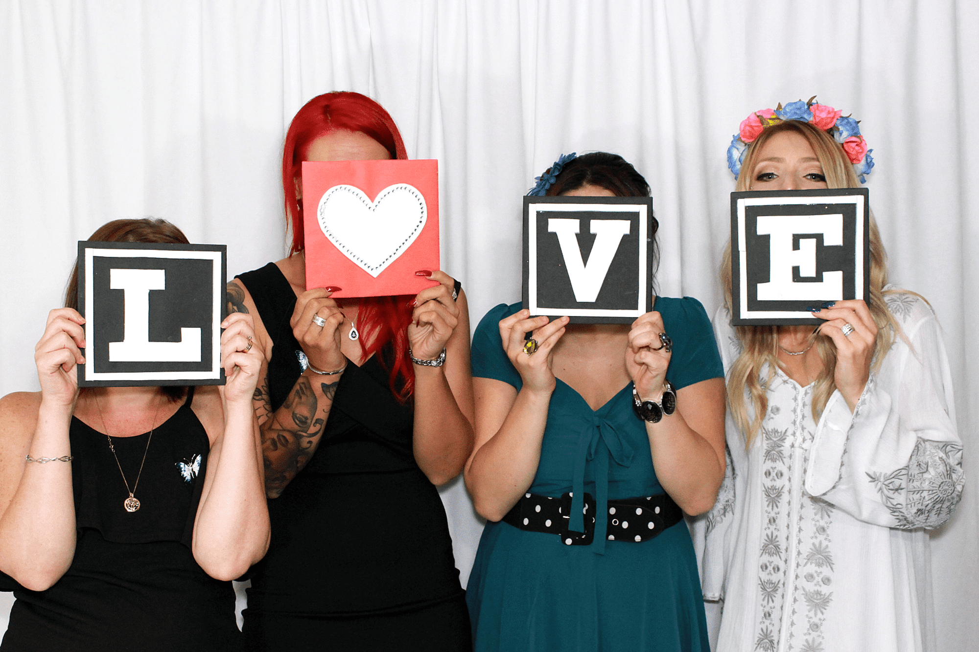 Four women in front of a white backdrop hold cards spelling LOVE for a playful wedding photo.