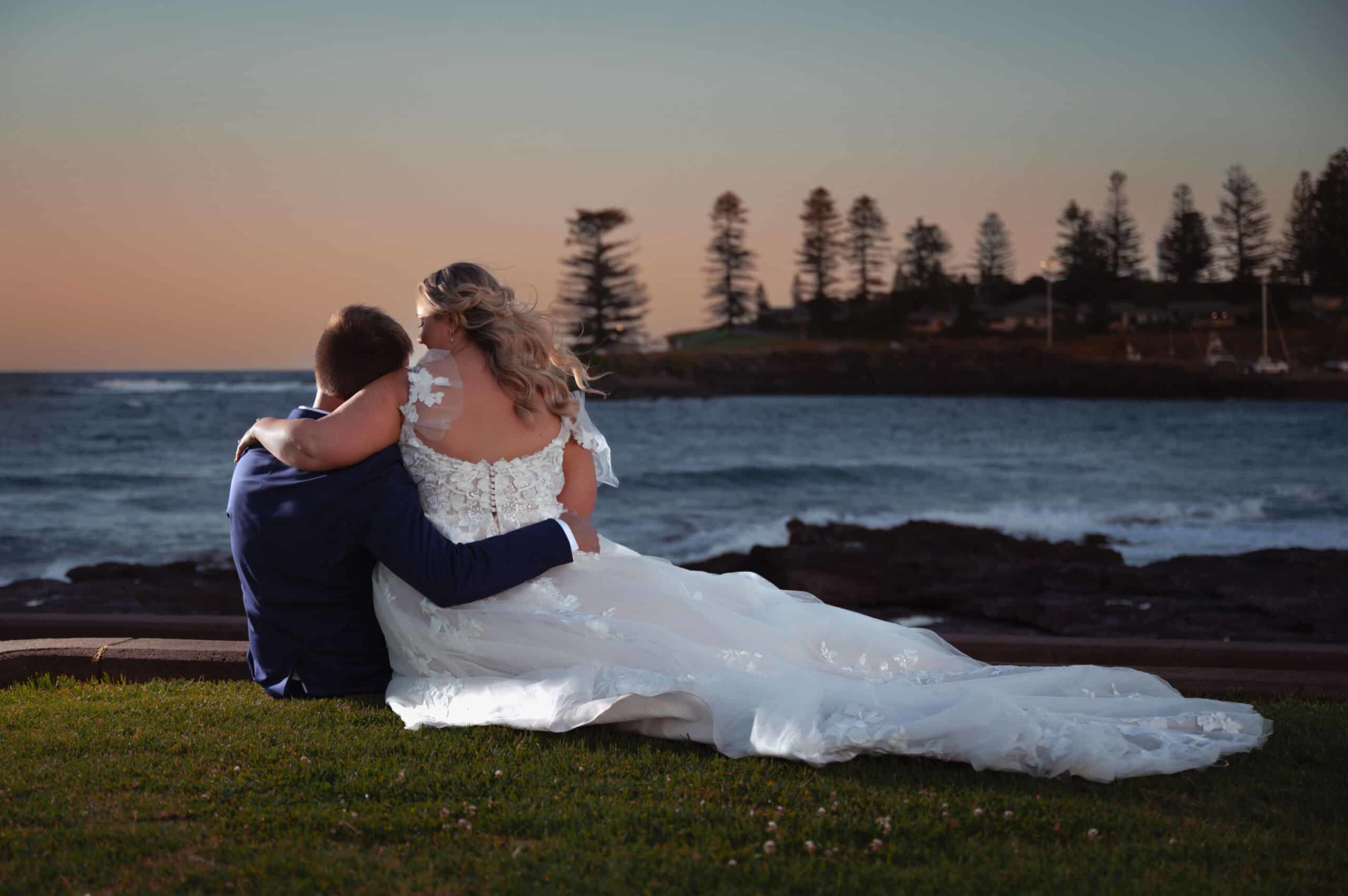 Bride and groom sit embracing on grass by the ocean at sunset, looking out over the water and distant trees.