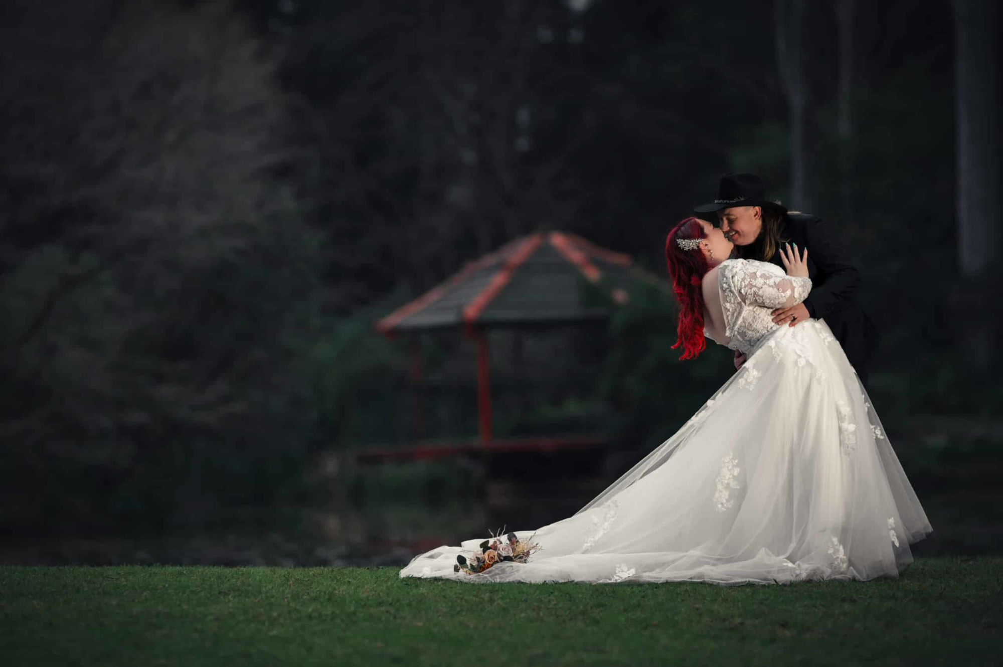 A bride in a flowing lace gown is dipped and kissed by her partner in a dark garden with a gazebo in the background.