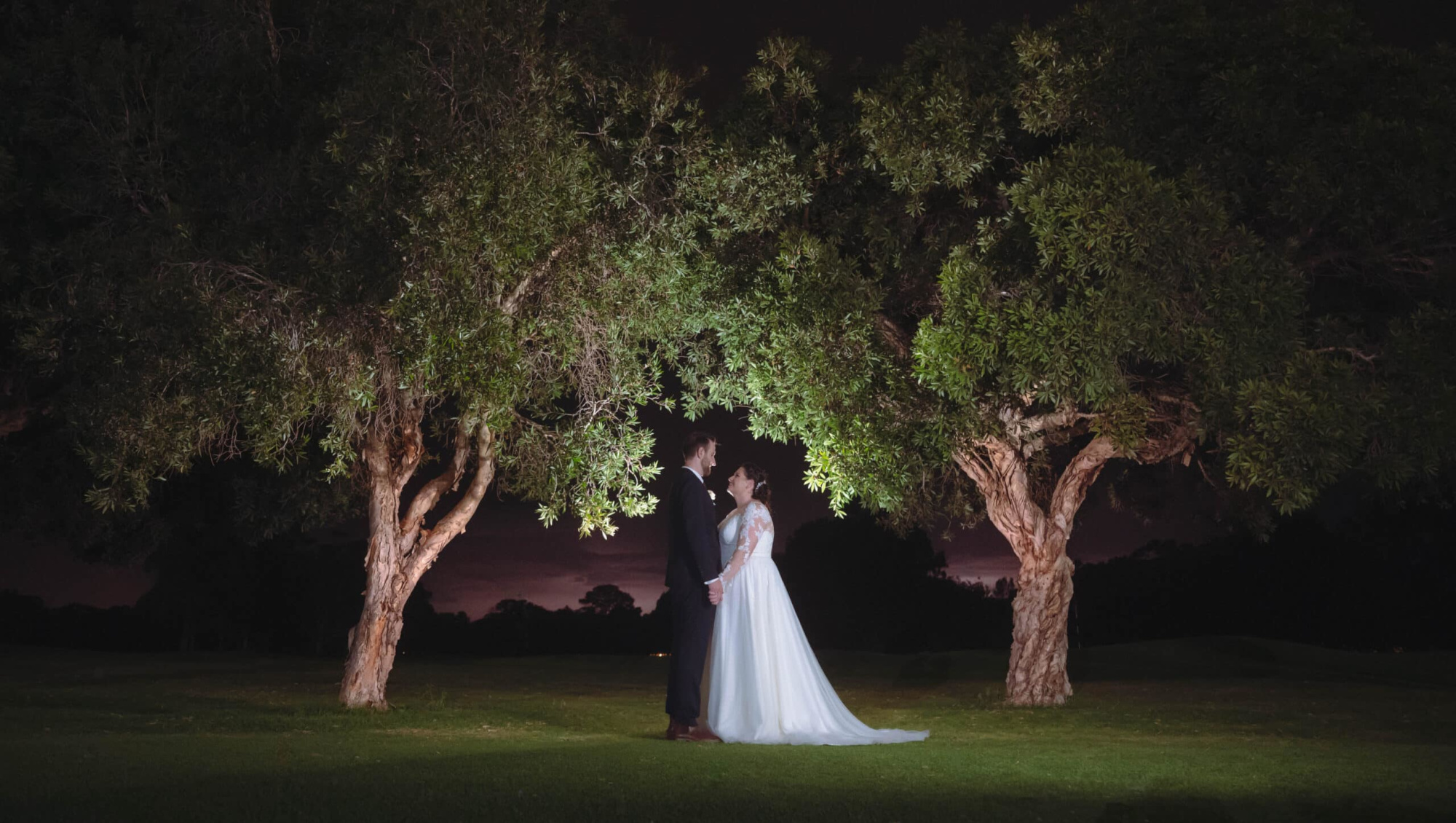 Bride and groom stand together under two illuminated trees at night on a grassy lawn.