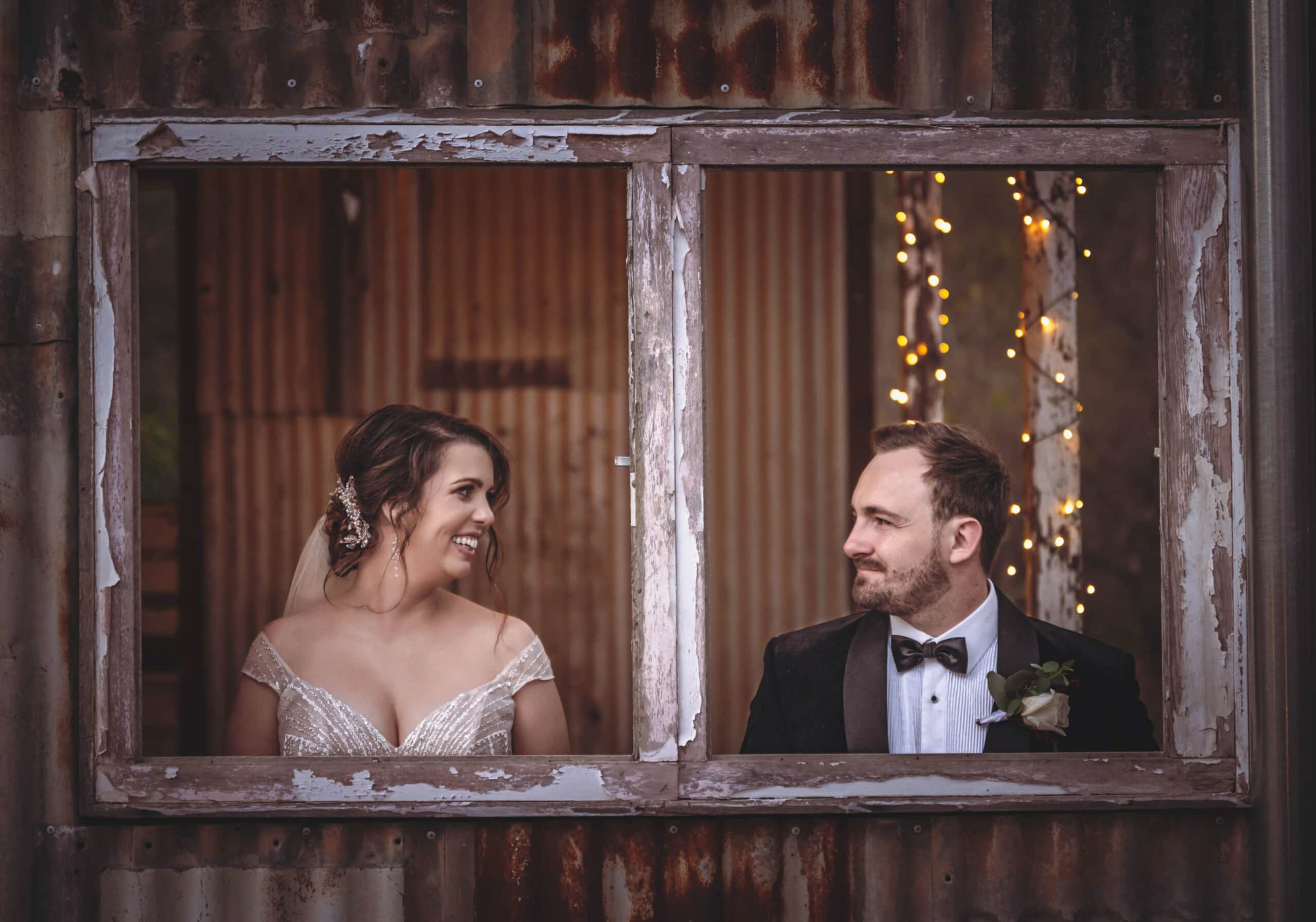 Bride and groom smile at each other through a rustic wooden window framed by warm fairy lights.