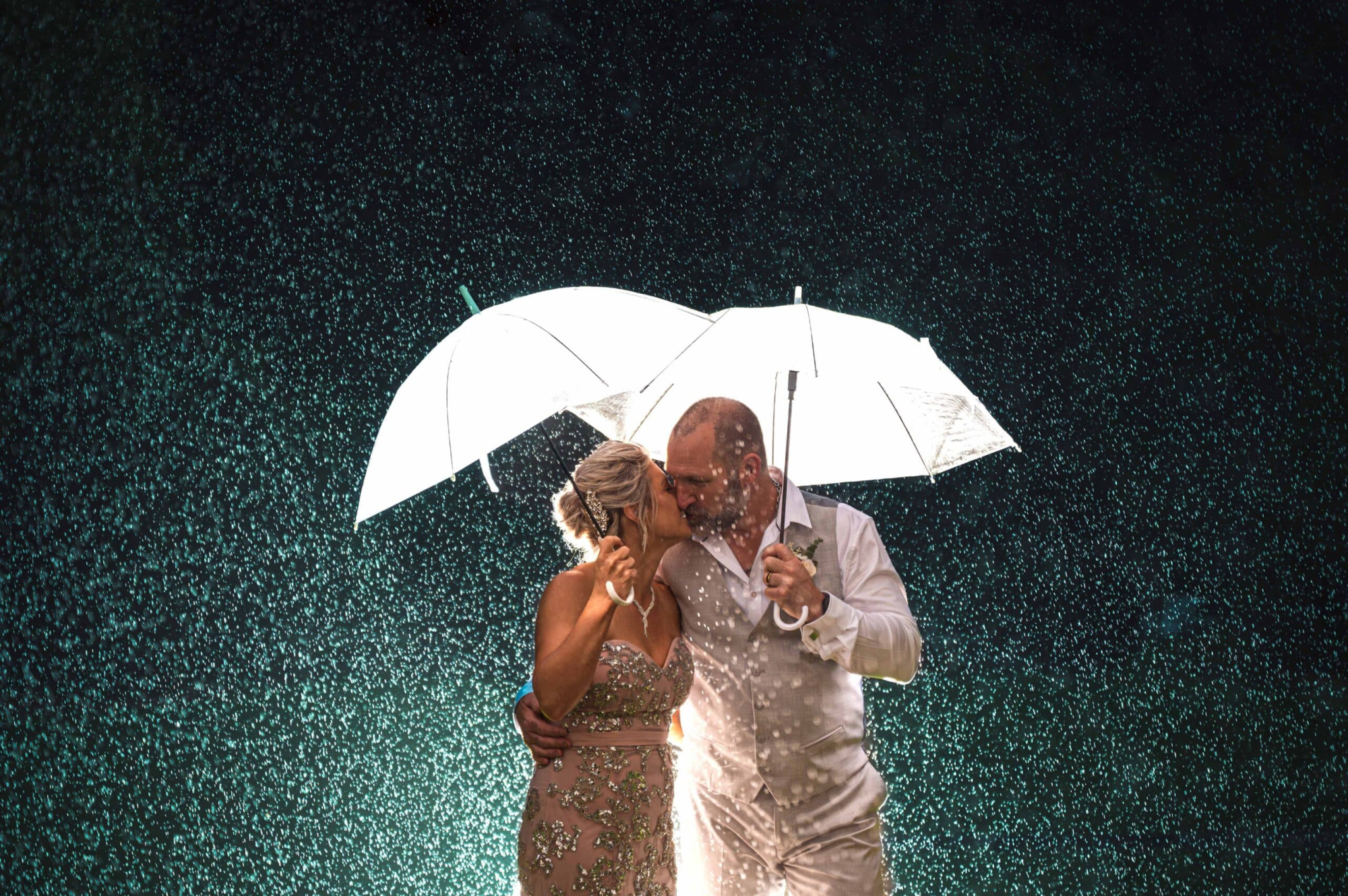 Bride and groom share a kiss under white umbrellas in the rain at night.