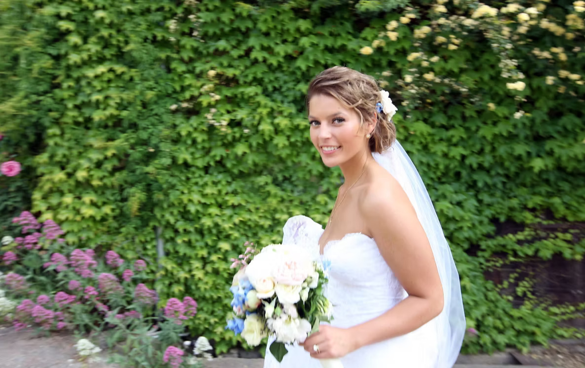 Bride in a strapless white gown holding a pastel bouquet in front of a lush green garden wall.