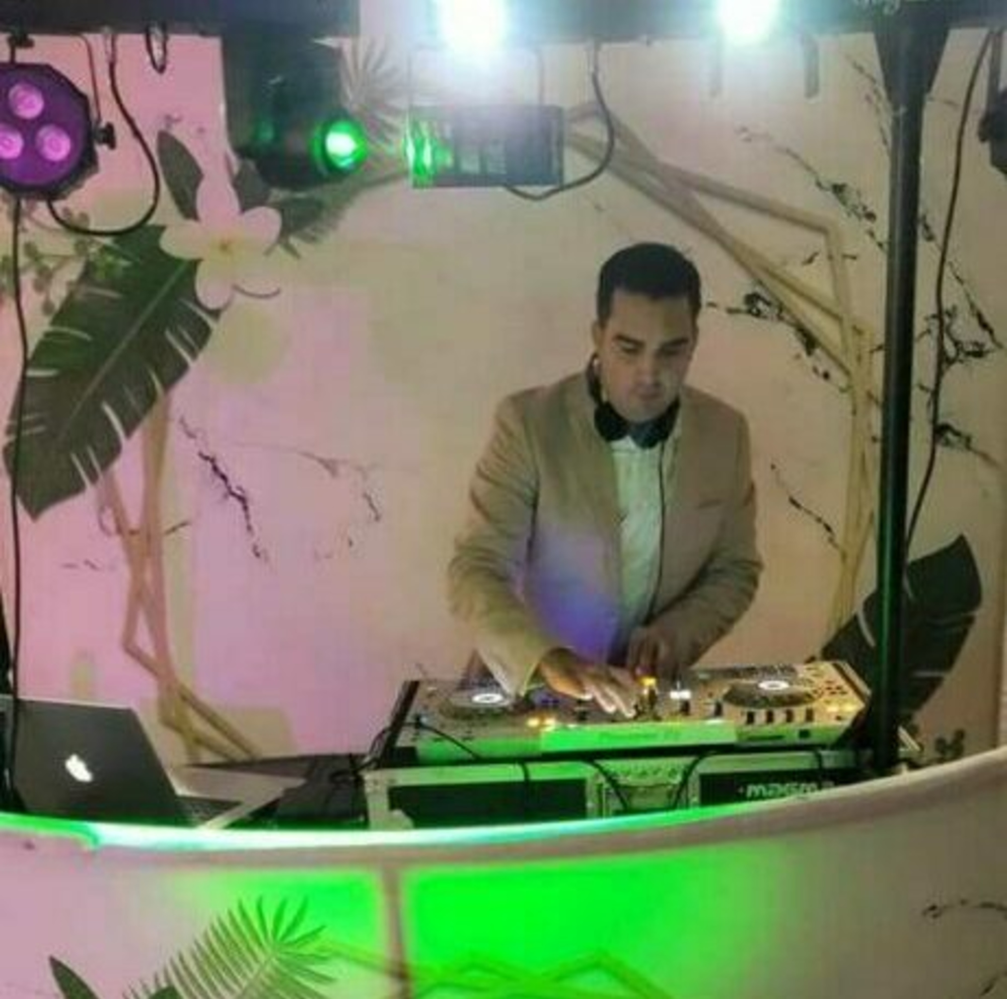 Wedding DJ mixing music at a modern booth with colorful lights and tropical backdrop.