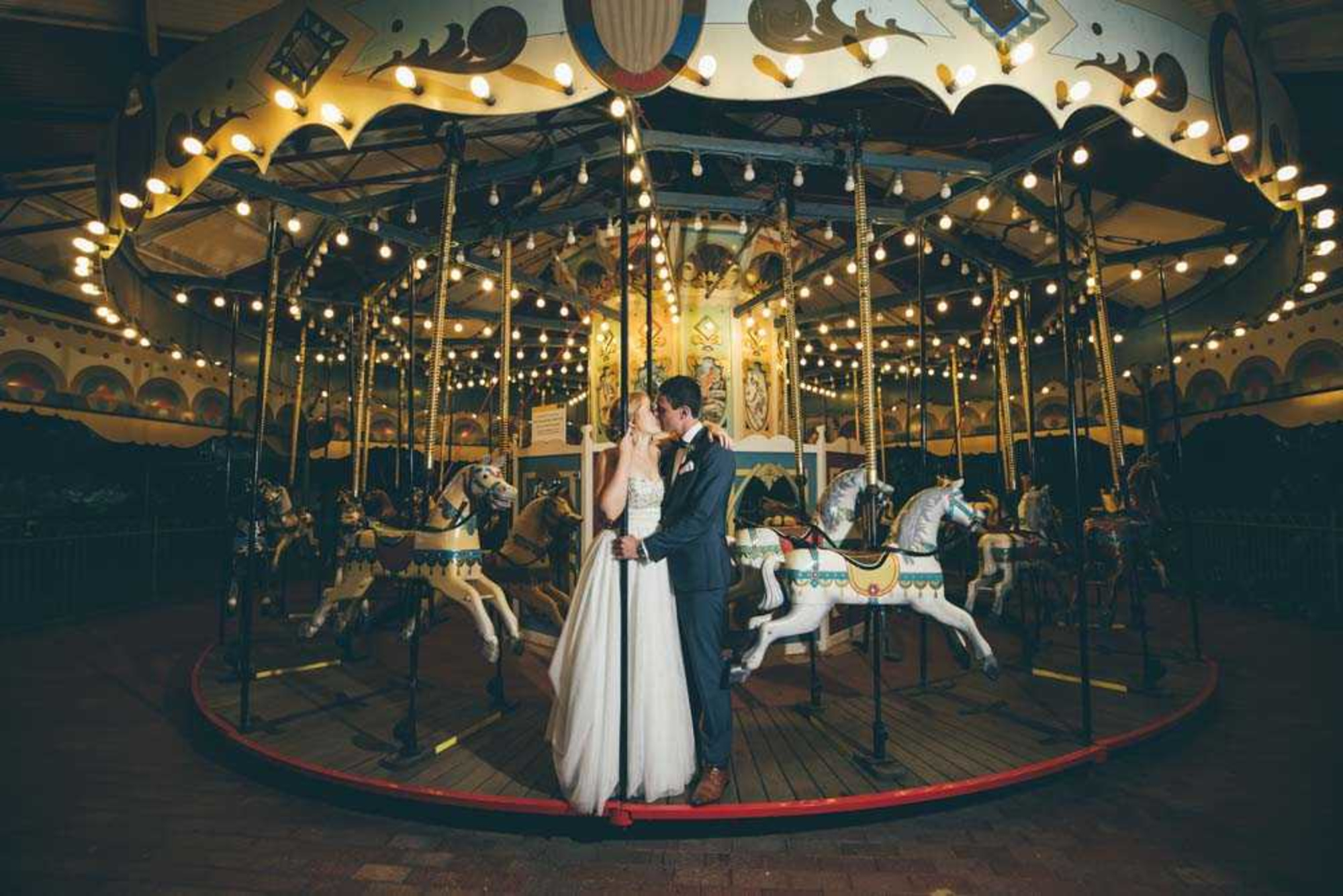 Bride and groom share a romantic kiss in front of a brightly lit carousel at night.