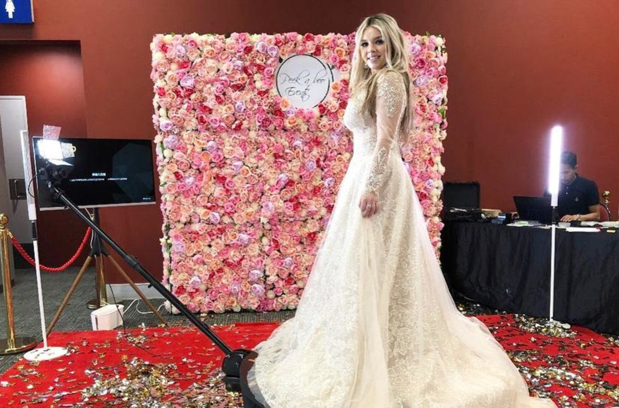 Bride in a long lace wedding gown posing on a red carpet in front of a pink flower wall photo booth setup.