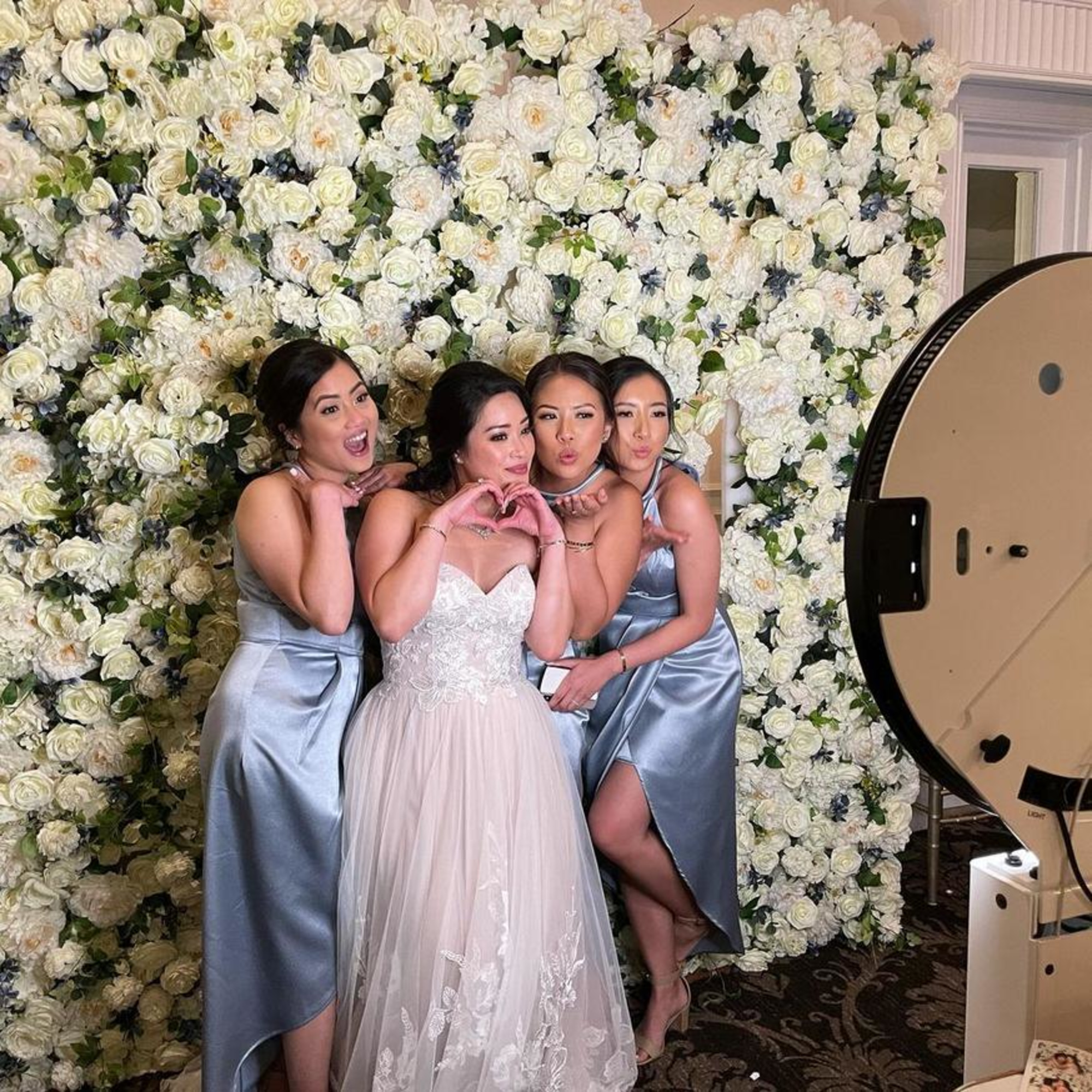 Bride and bridesmaids pose at a wedding photo booth in front of a white flower wall backdrop.