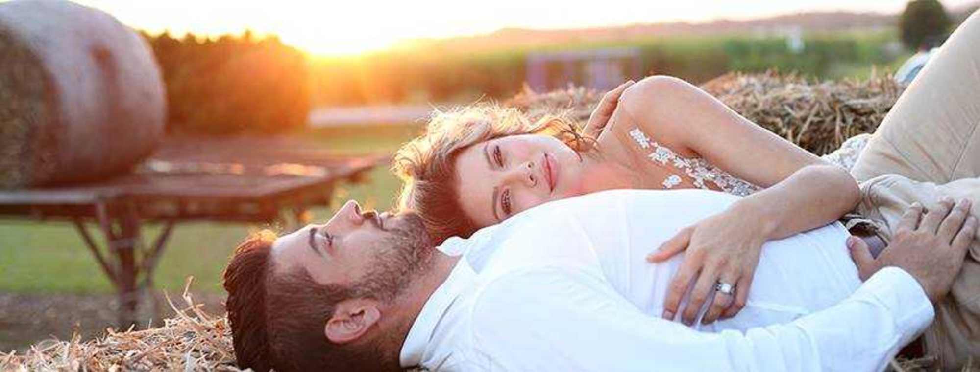 A couple in wedding attire cuddles on hay bales at sunset in a rustic outdoor setting.