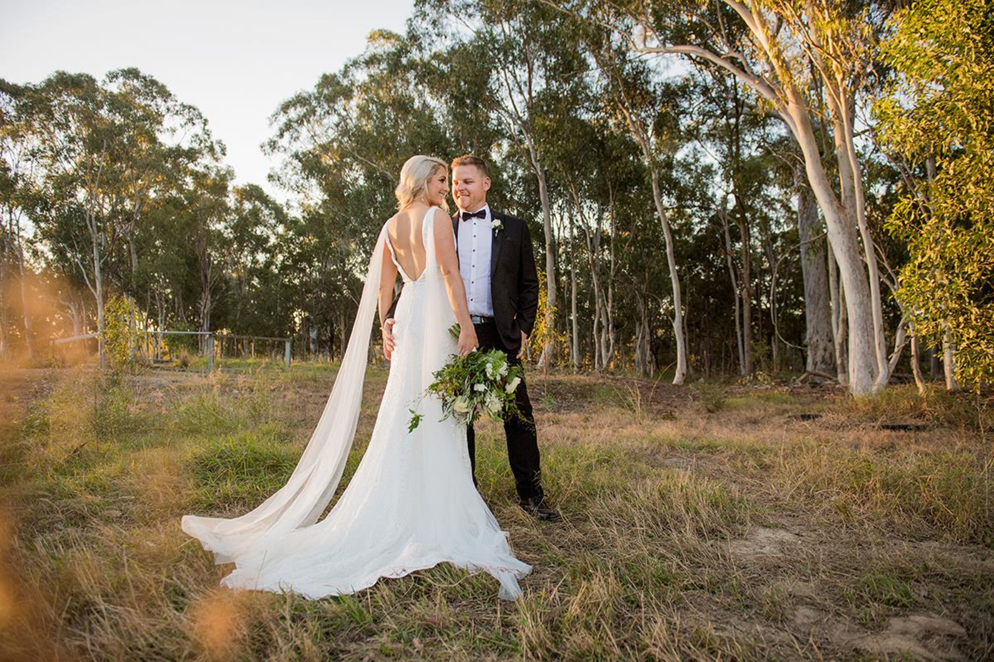 Bride and groom share a moment in a sunlit forest clearing, with her long train and bouquet trailing on the grass.