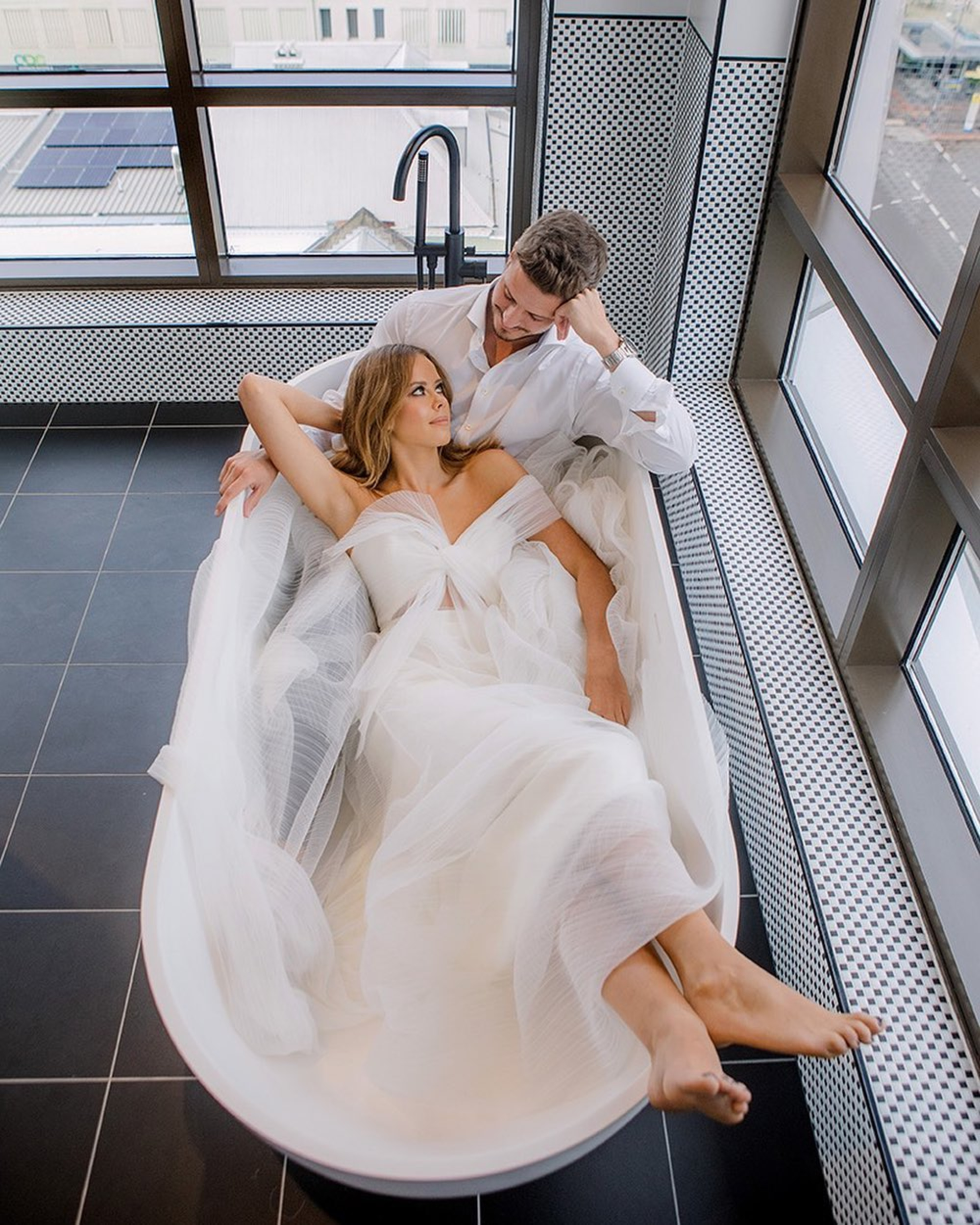 A bride in a flowing white gown lounges in a bathtub with her partner in a modern city loft bathroom.
