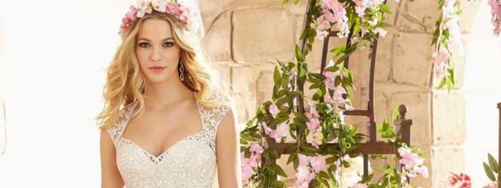 Bride in a lace wedding gown and flower crown standing by pink floral decorations in a stone courtyard.