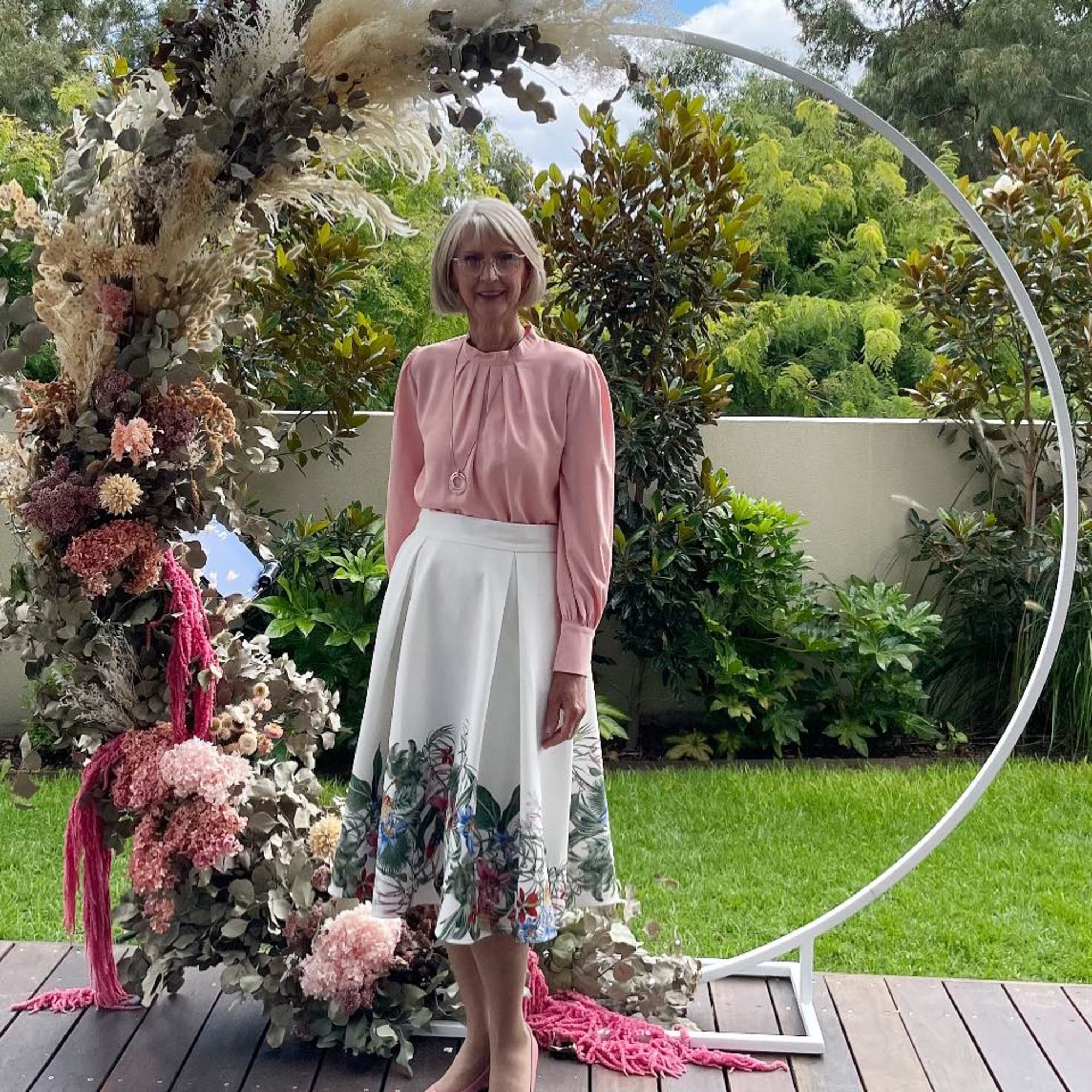 Female officiant stands by a circular floral wedding arch in a lush garden setting.