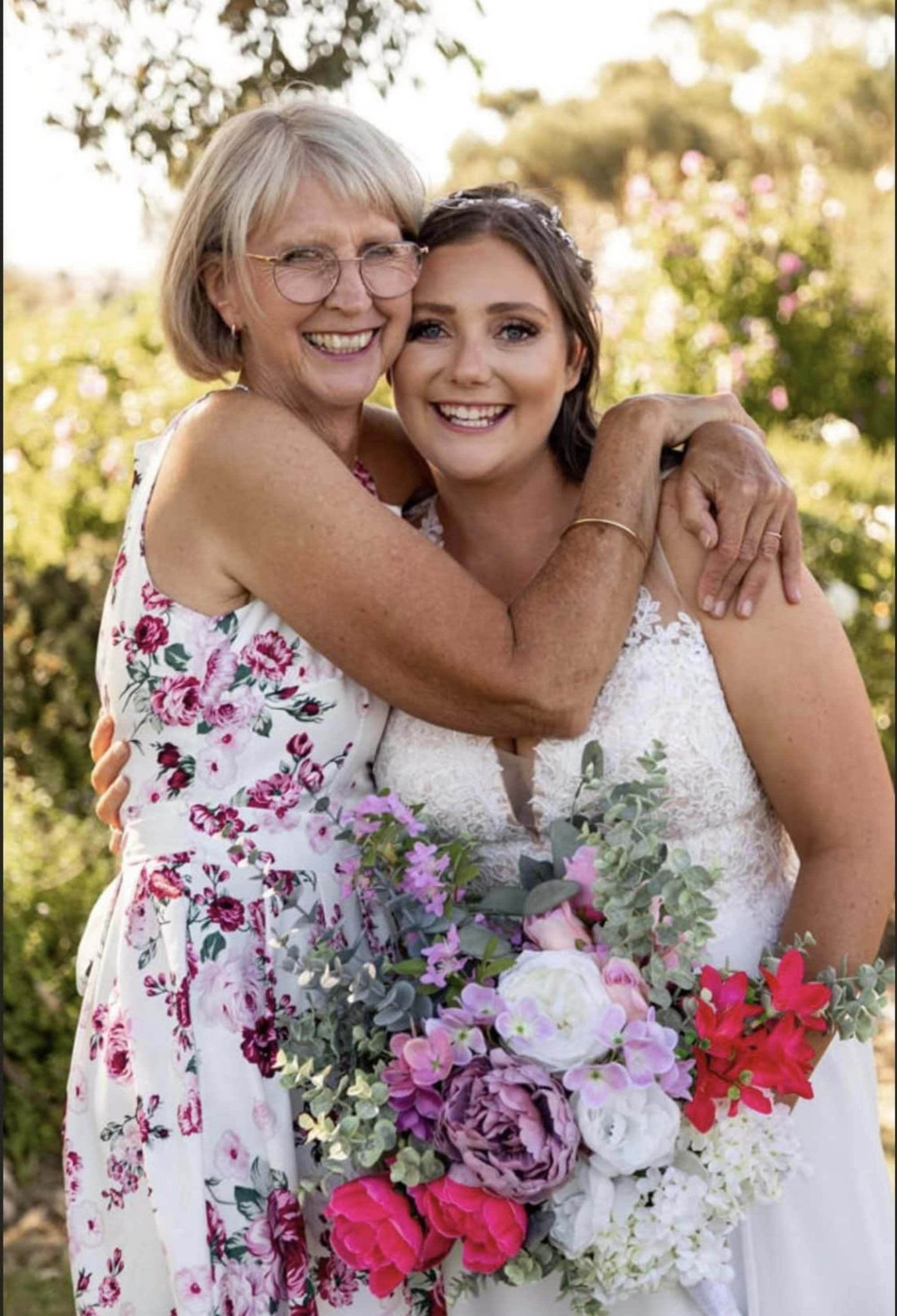 Bride and older woman hugging and smiling outdoors, holding a colorful floral wedding bouquet.