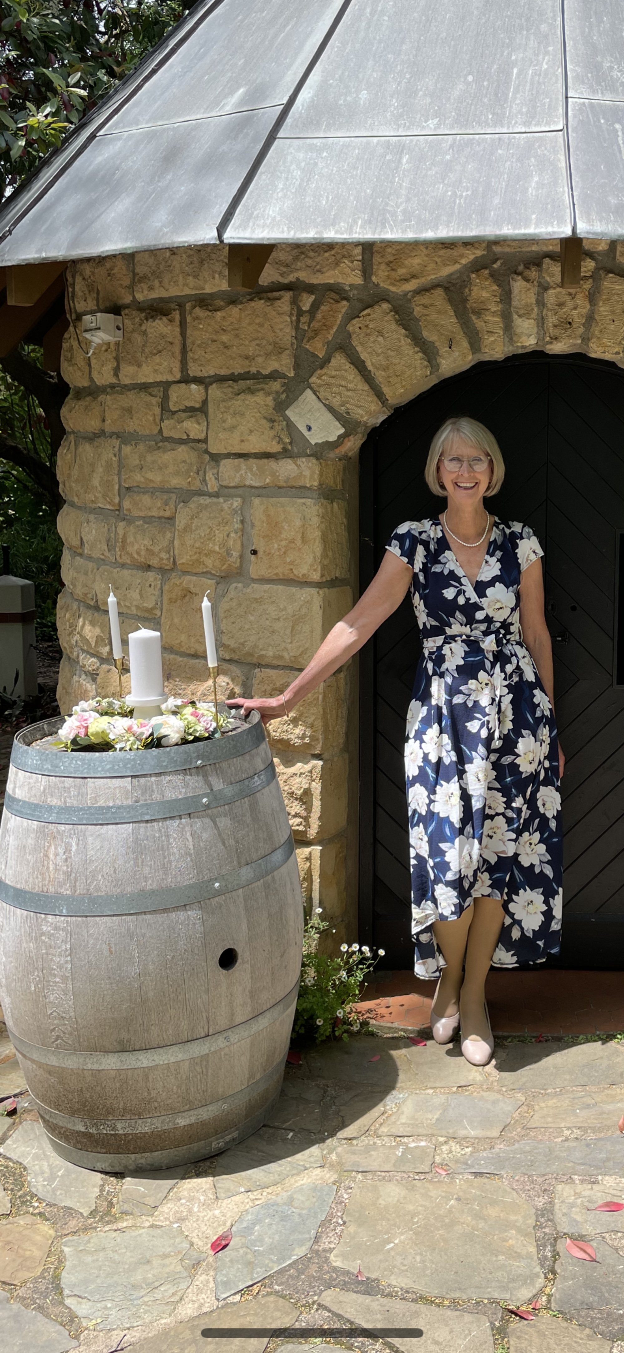 Smiling wedding celebrant standing by a flower-topped wine barrel with candles at a rustic stone venue entrance.