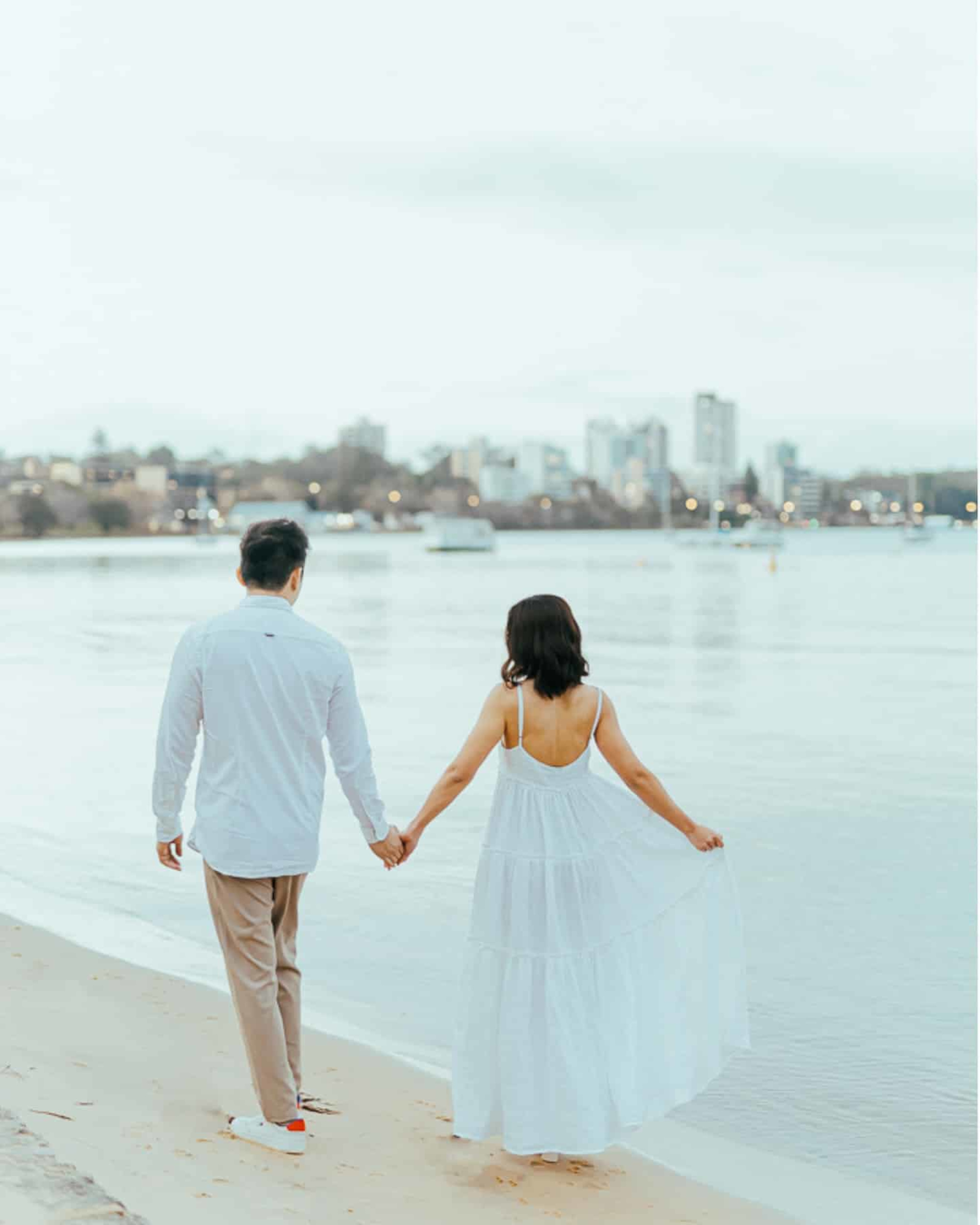 A couple holding hands while walking along a calm waterfront beach with a city skyline in the background.