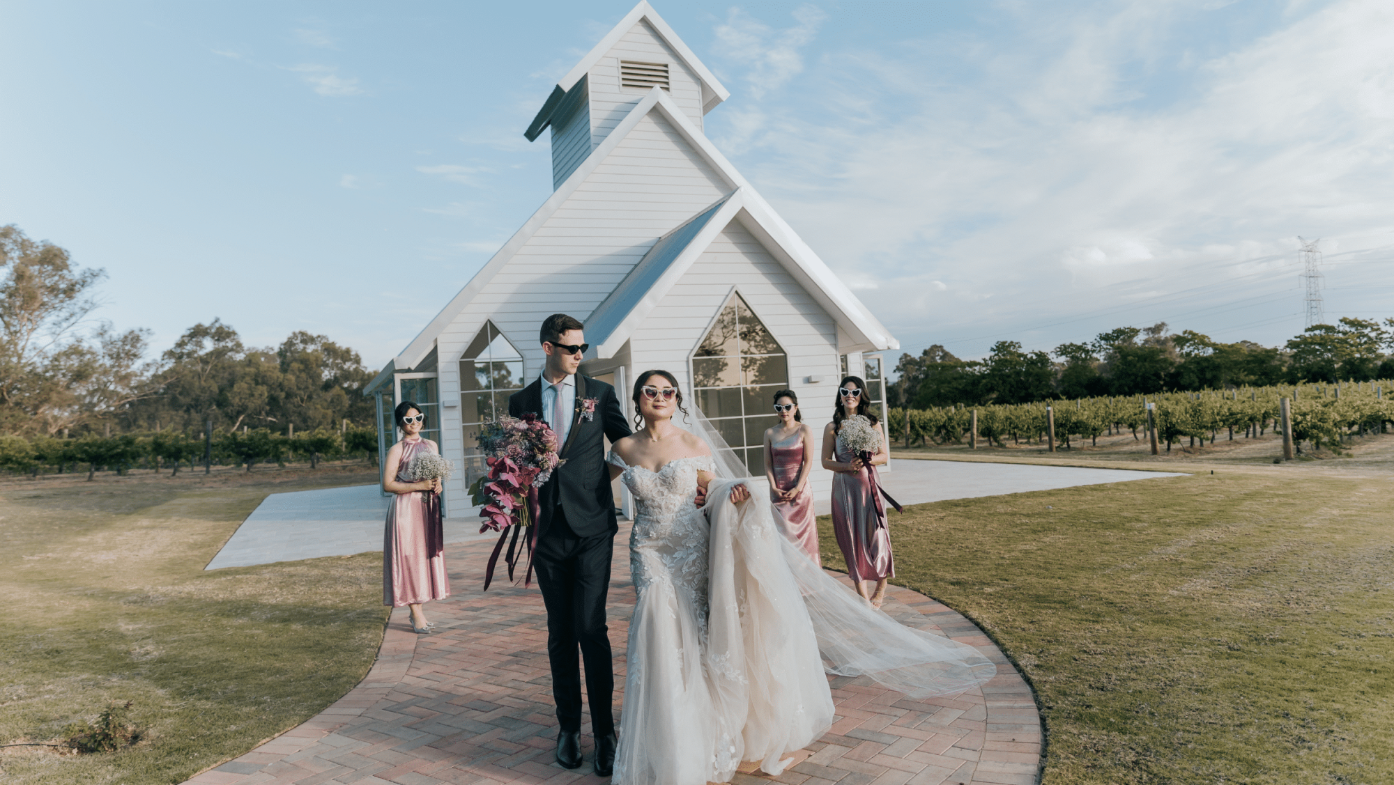 Bride and groom in sunglasses walk from a white chapel with their bridesmaids at a vineyard wedding.