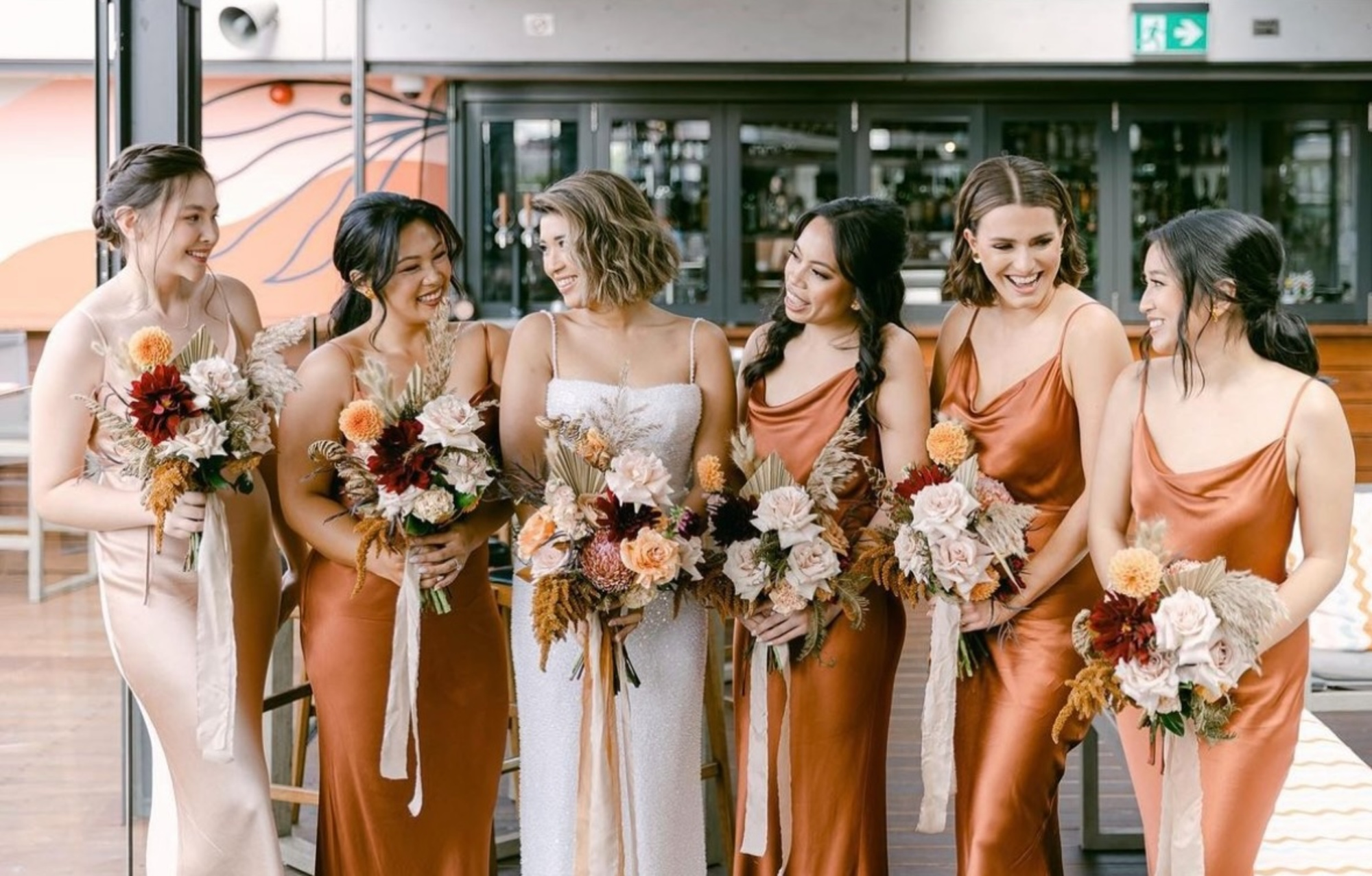 Bride and bridesmaids in rust satin dresses holding autumn-toned bouquets and laughing together indoors.