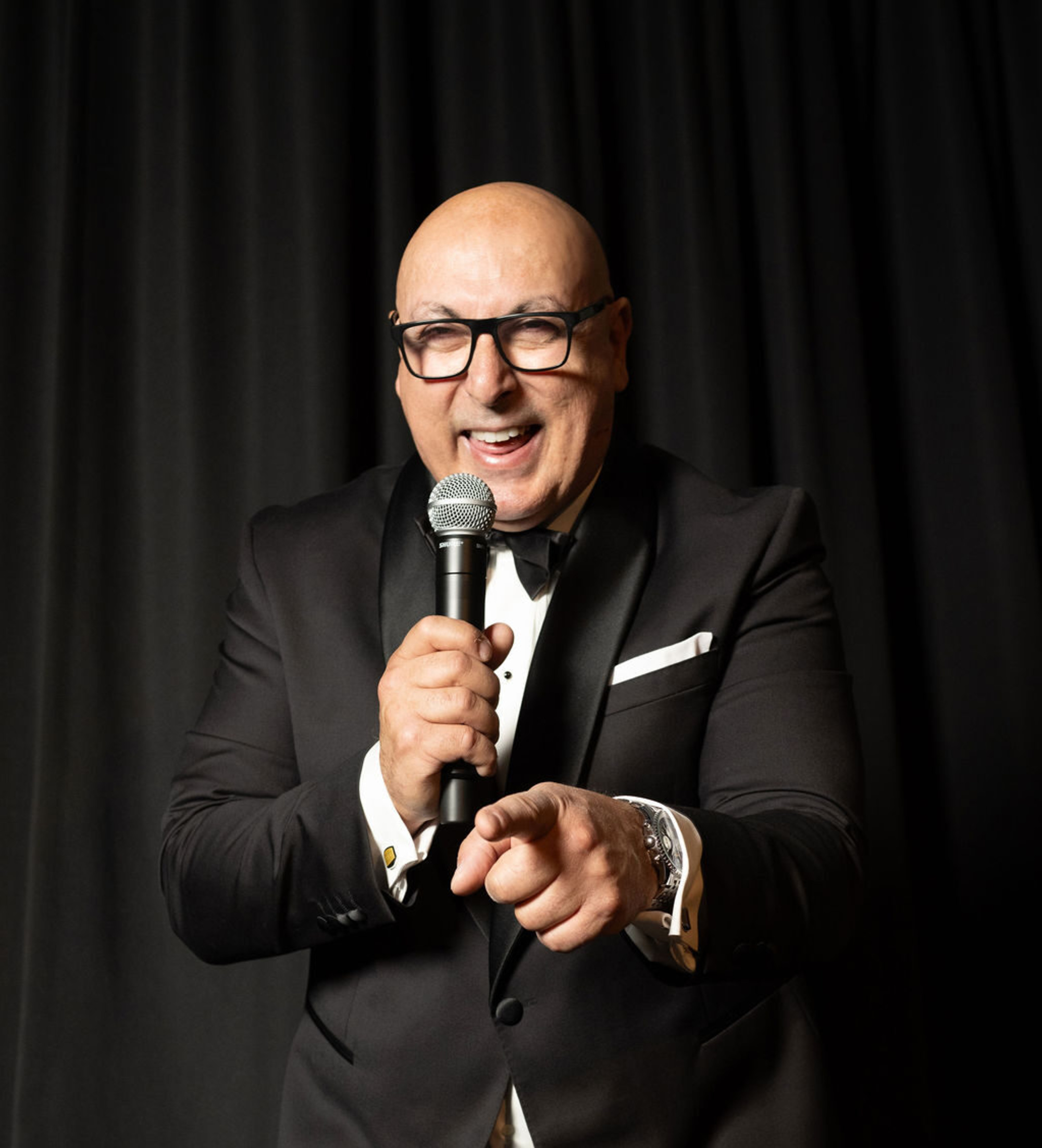 Smiling wedding emcee in a black tuxedo holding a microphone and pointing toward the camera against a dark backdrop.
