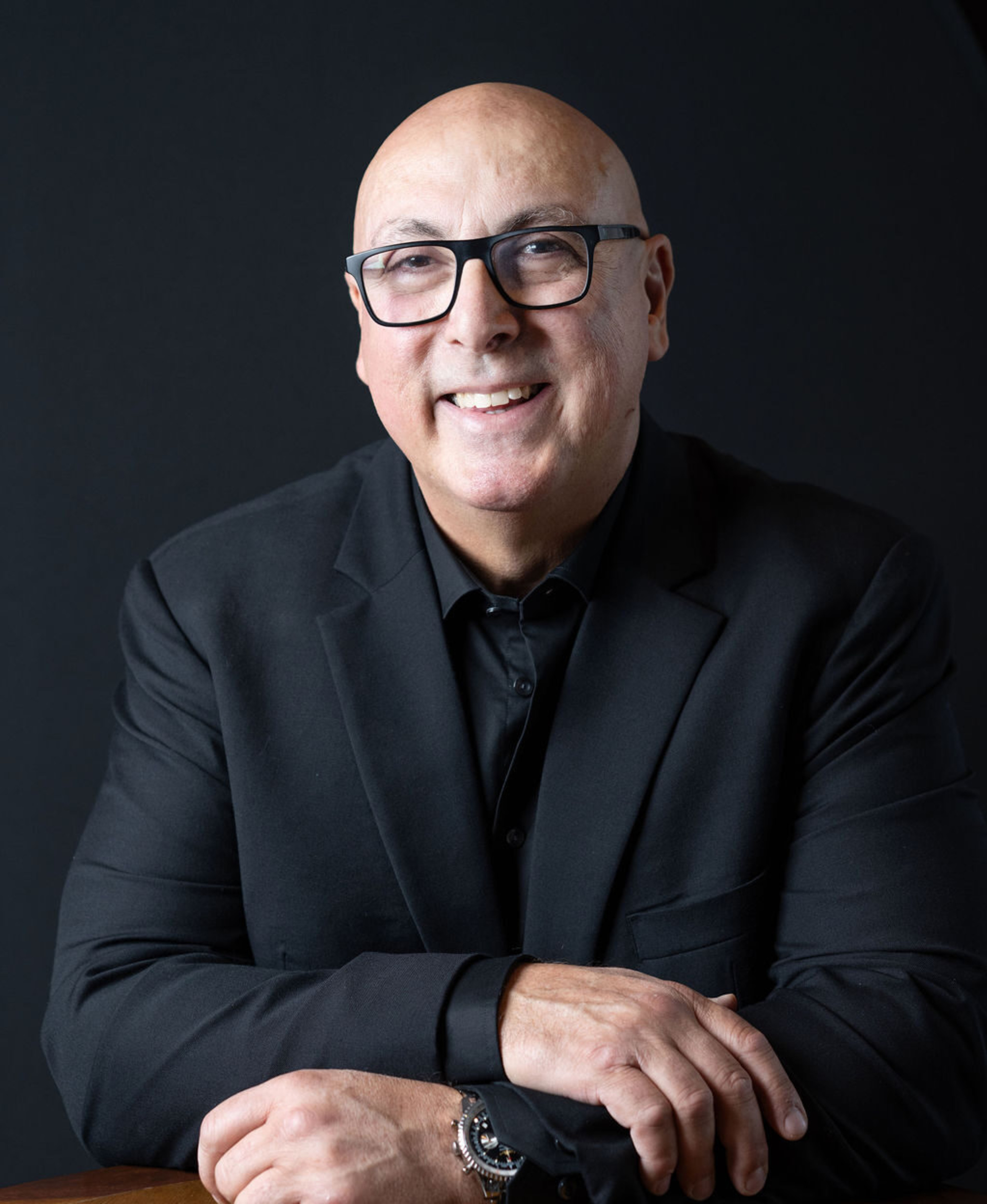 Smiling man in a black suit and glasses posing for a professional headshot against a dark background