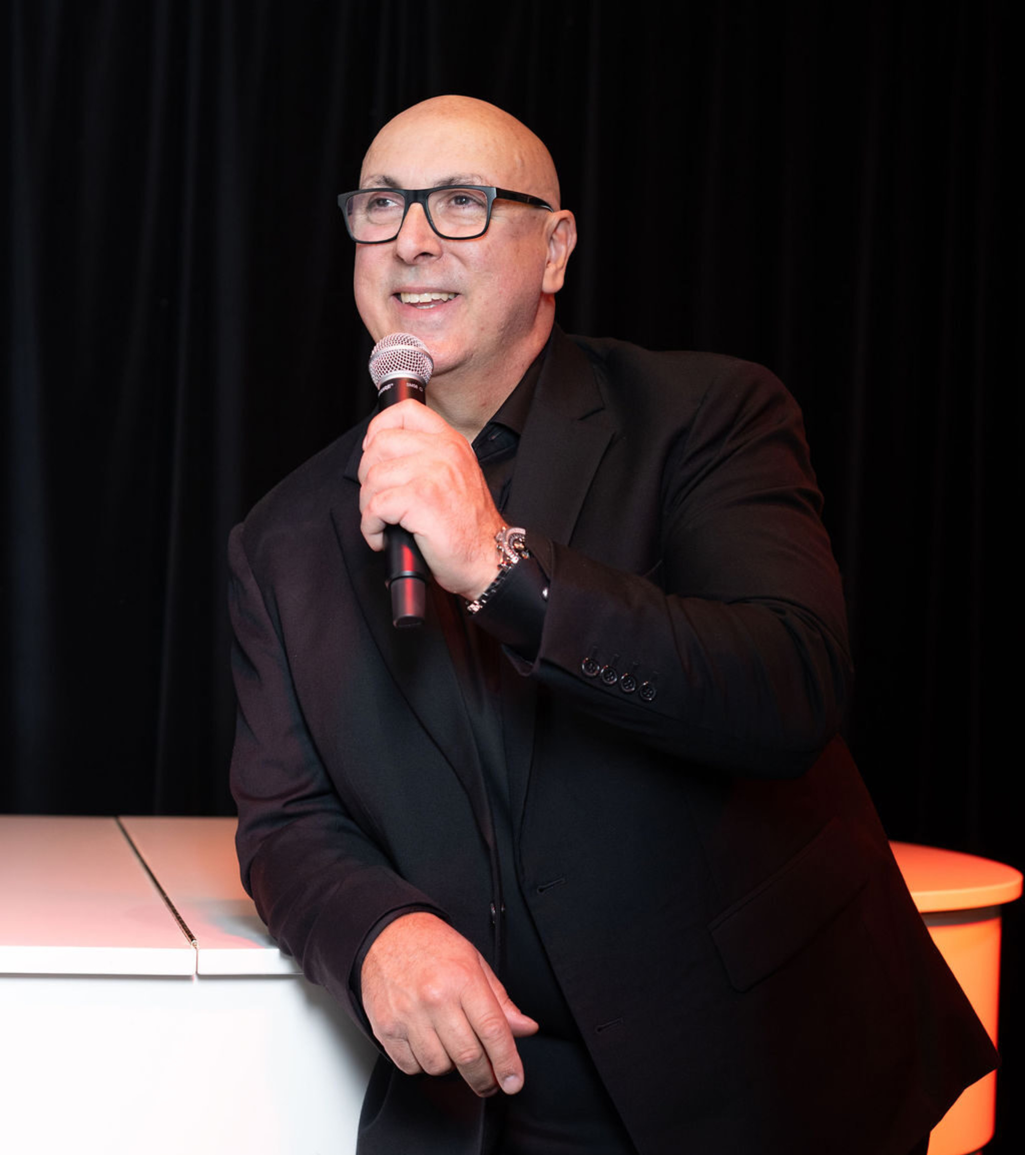 Wedding emcee in a black suit speaking into a microphone against a dark backdrop.