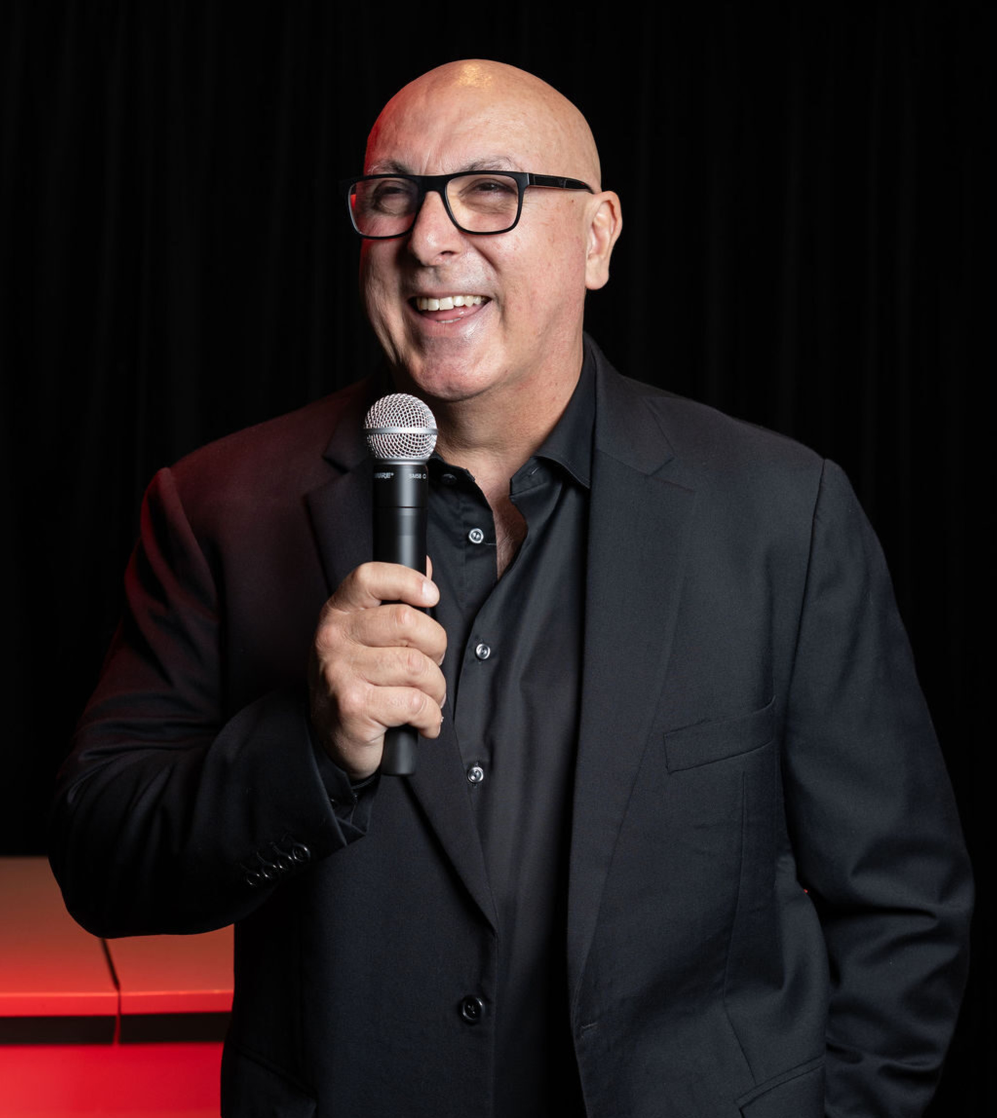 Smiling man in a dark suit holding a microphone against a black backdrop, posed like a wedding emcee.