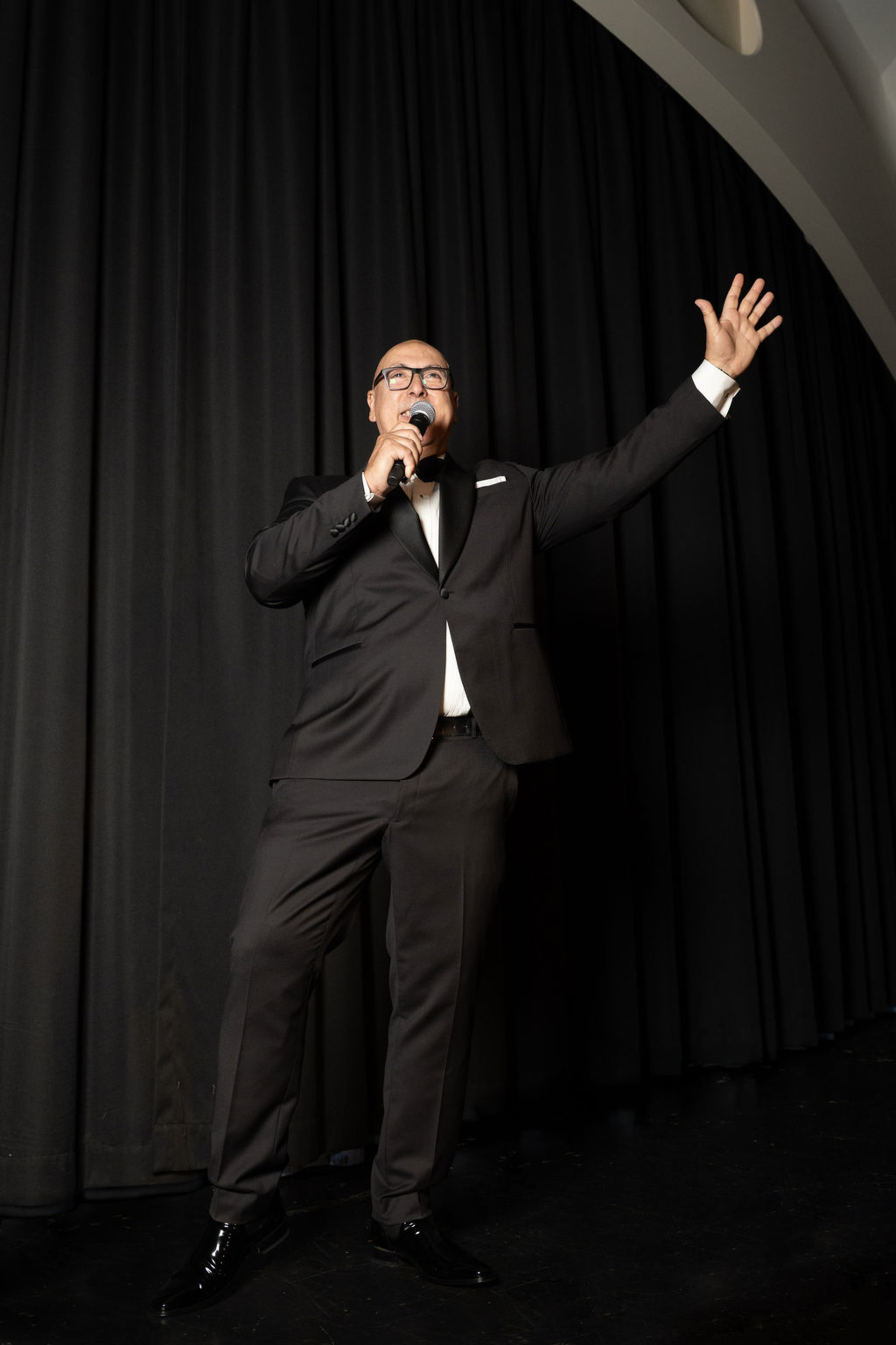 Wedding emcee in a tuxedo speaking into a microphone on stage with black curtains behind him.