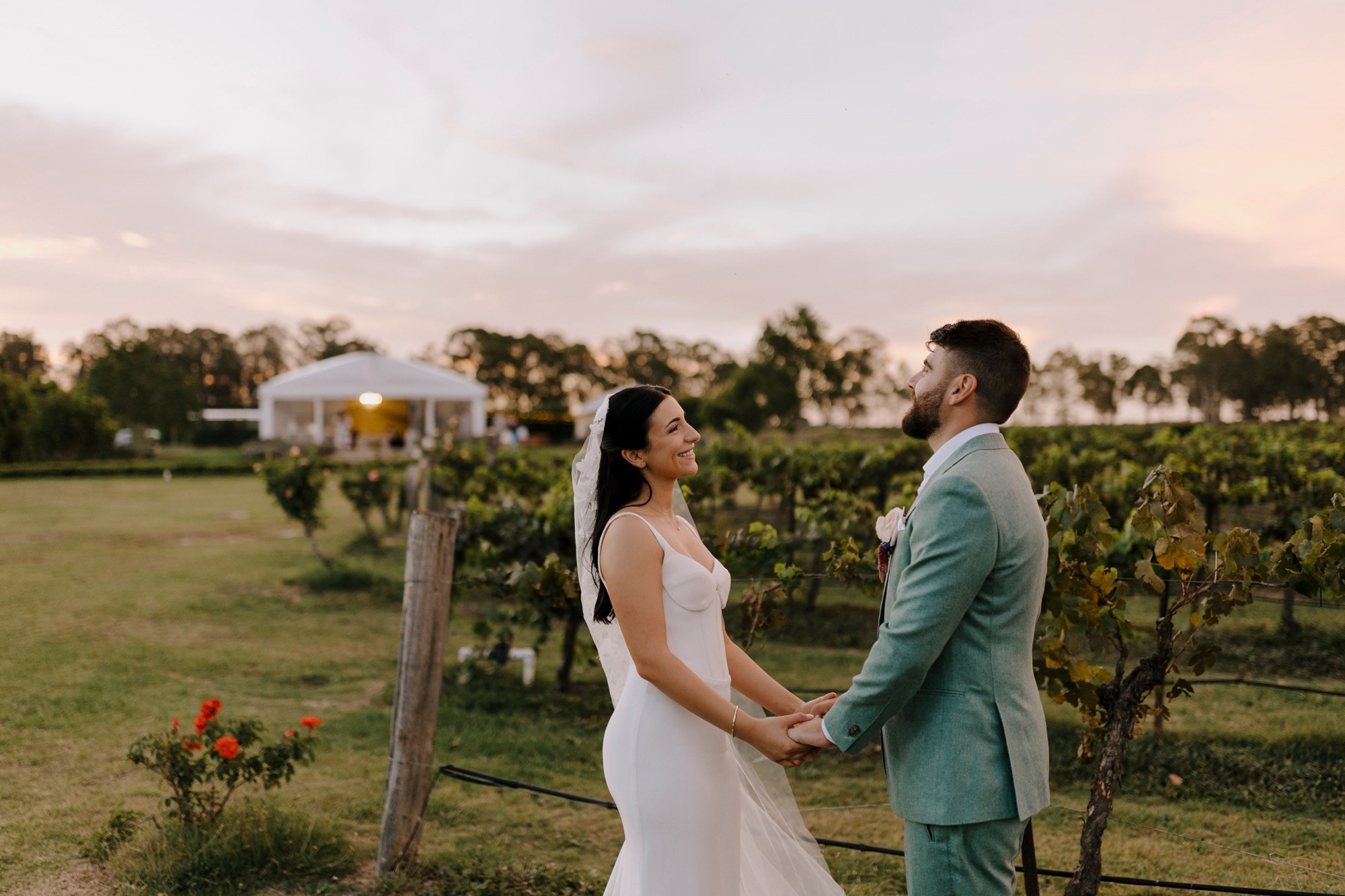 Bride and groom hold hands in a vineyard at sunset with a white marquee in the background.