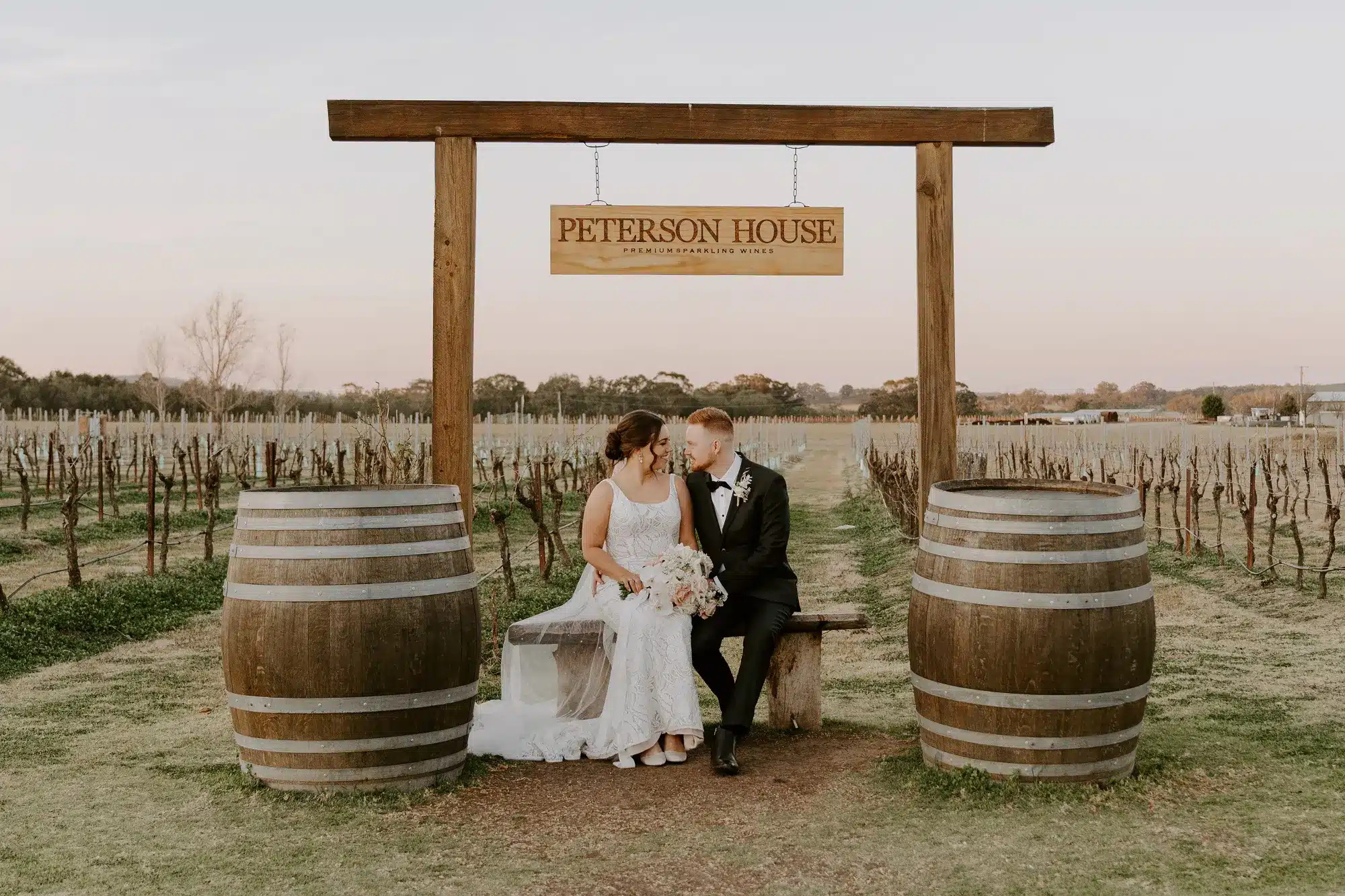 Bride and groom sit beneath a wooden Peterson House sign in a rustic vineyard framed by wine barrels at sunset.