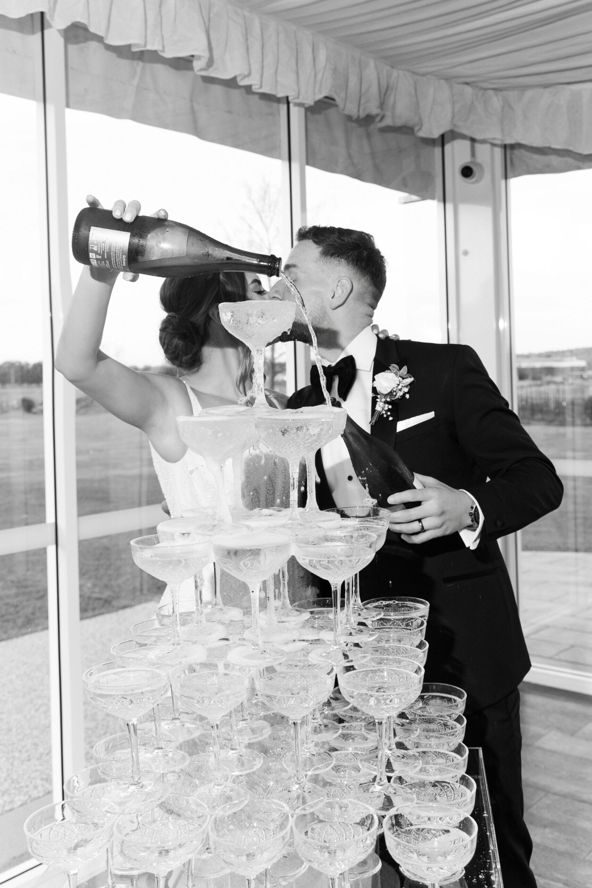 Bride and groom share a kiss while pouring champagne into a tower of coupe glasses at their wedding reception.