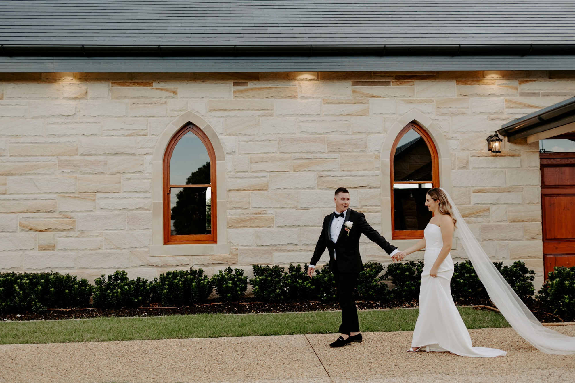 Bride and groom walk hand in hand outside a stone chapel with arched wooden windows.