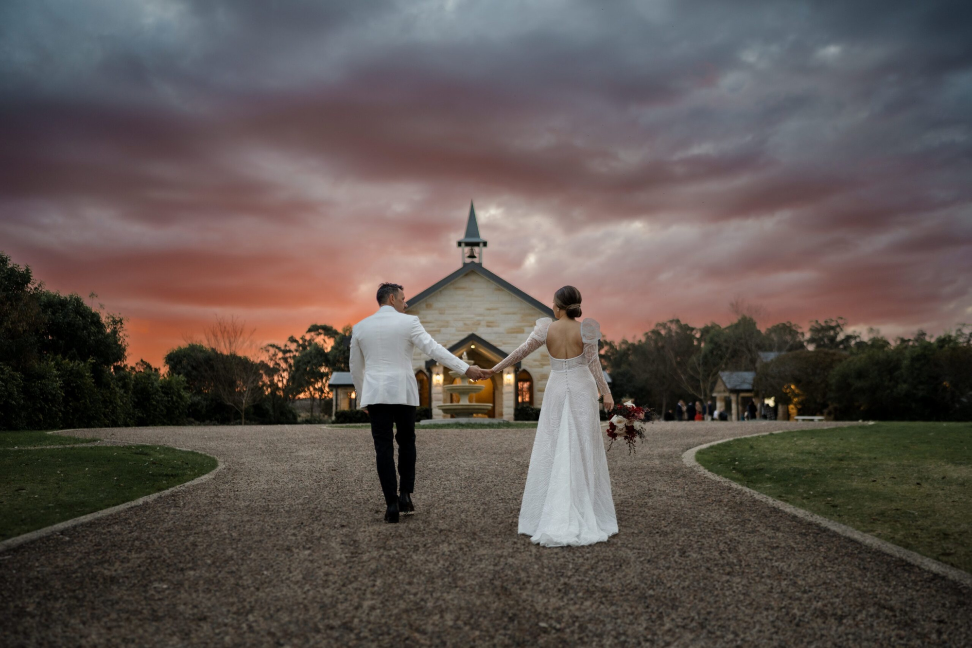 Couple walks hand in hand toward a small chapel at sunset under a dramatic sky.